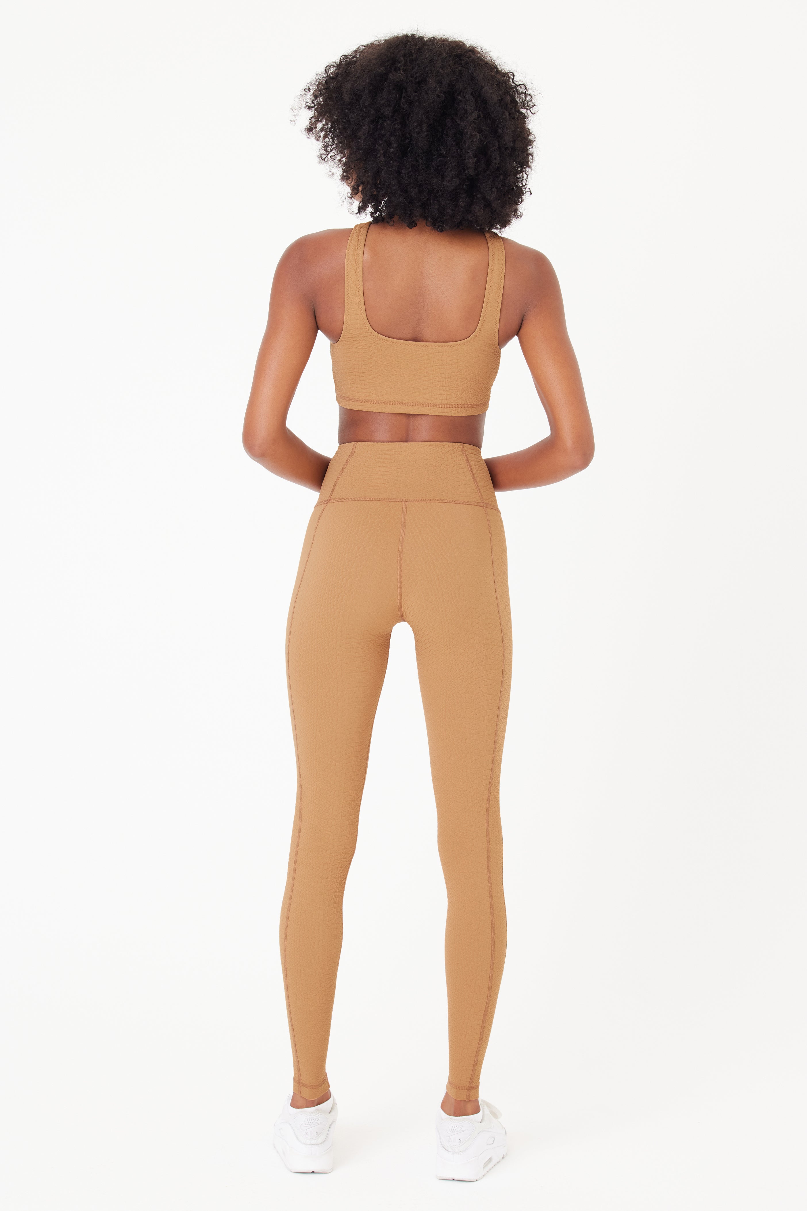A curly-haired person models a tan-colored set, featuring a high-waist Ava Legging by Gigi C Sport, made from compression waffle jacquard fabric, with their back to the camera. They wear white sneakers against a plain white background.