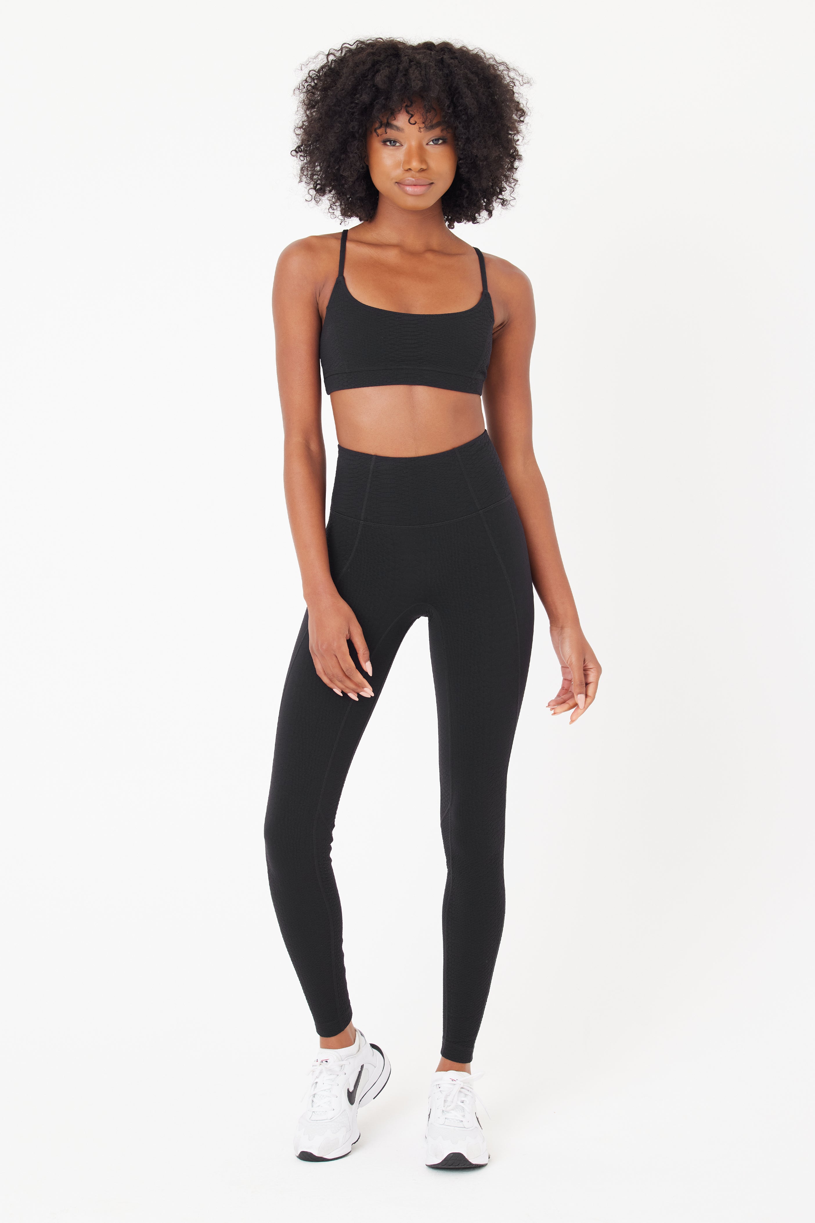 The curly-haired woman confidently sports a Gigi C Sport Ava Legging set in bi-stretch compression waffle jacquard fabric with a black sports bra. She smiles brightly with white sneakers in a plain white background.