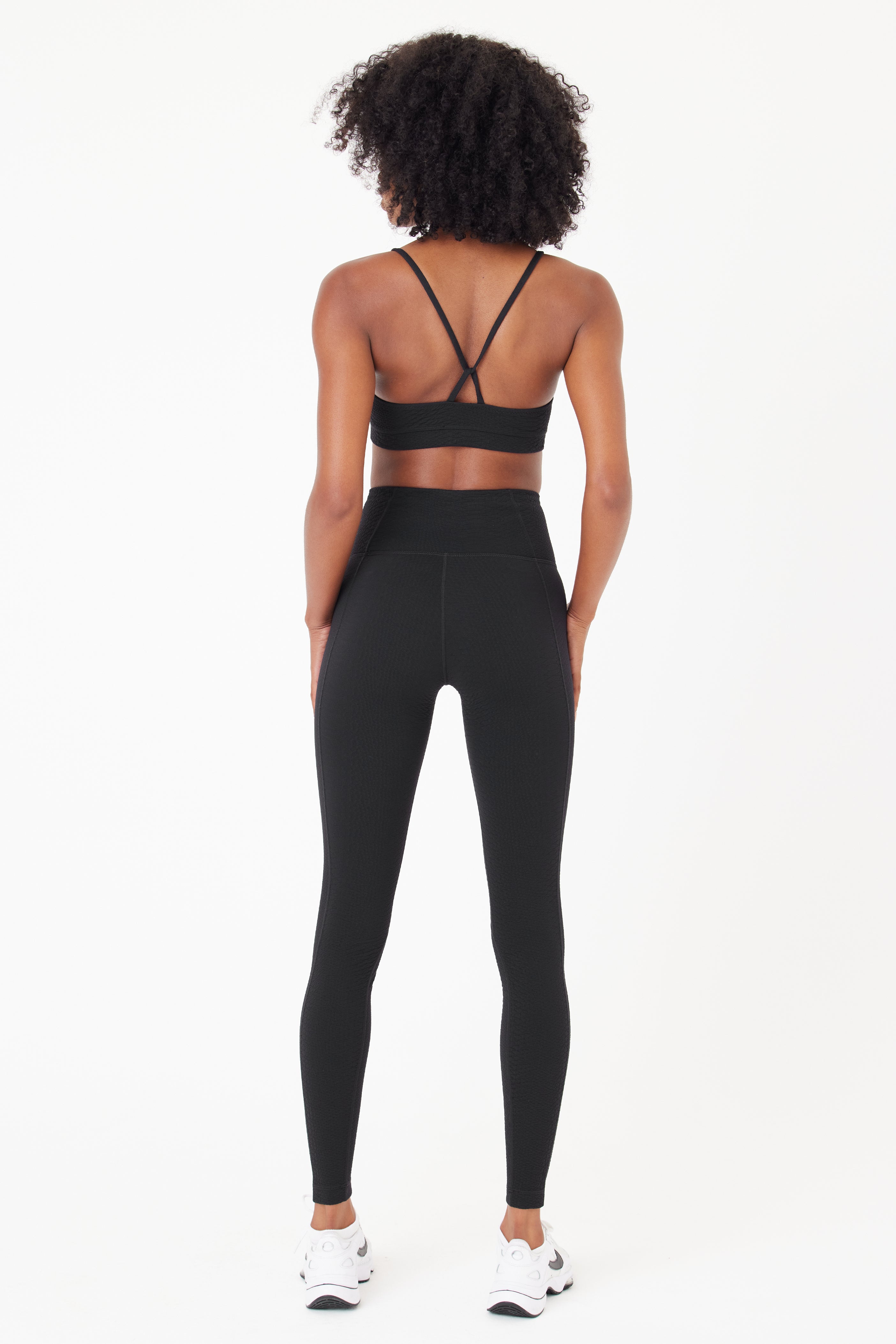 The curly-haired individual, wearing a black sports bra and Gigi C Sports Ava Legging in bi-stretch compression waffle jacquard, stands back to the camera. They complete their look with white sneakers against a plain white backdrop.