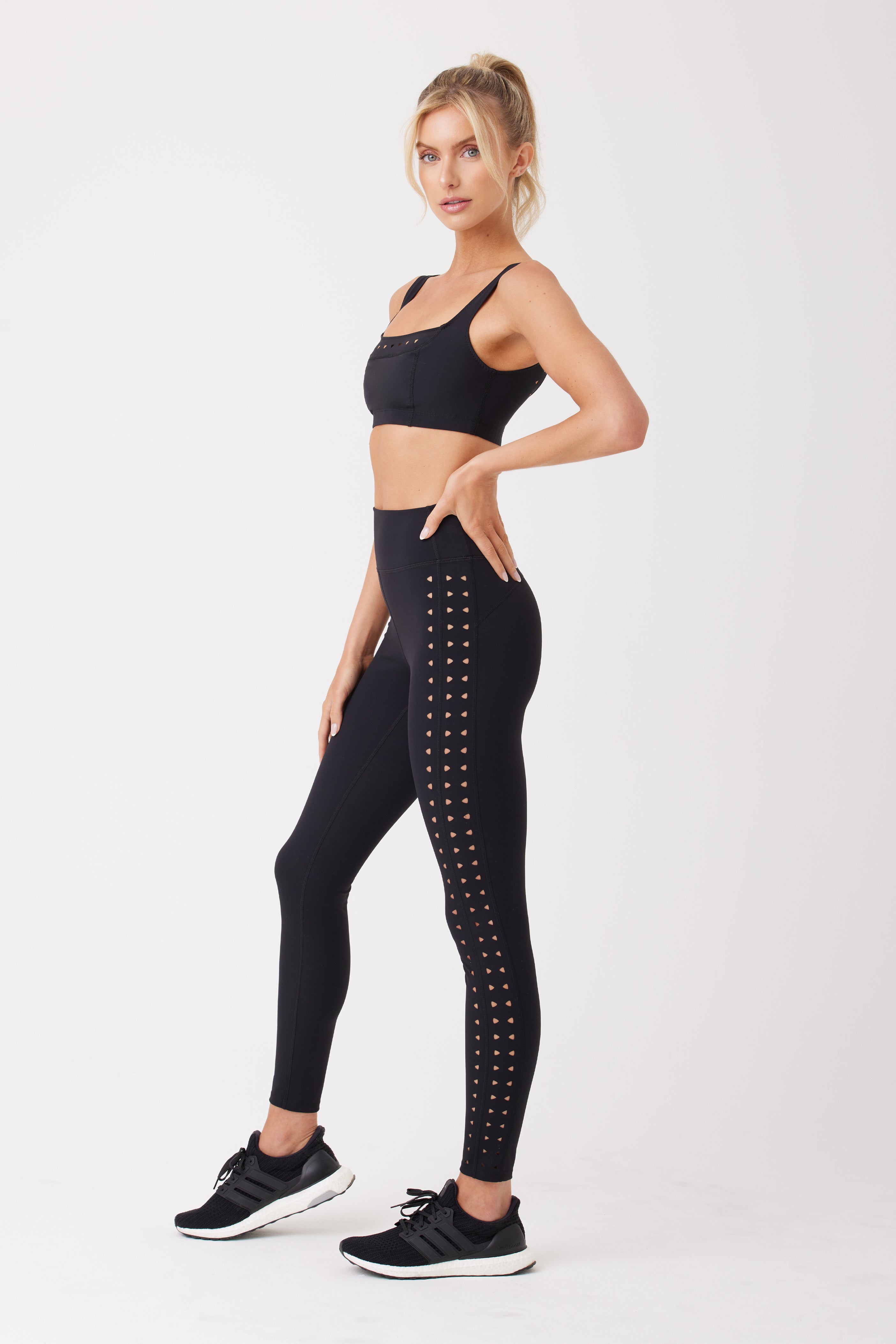 A woman poses against a white background in a black sports bra and Gigi C Sports Katrina Legging, featuring patterned side designs. Shes wearing black athletic shoes, her hair tied in a ponytail, highlighting the sleek style enhanced by compression fabric.