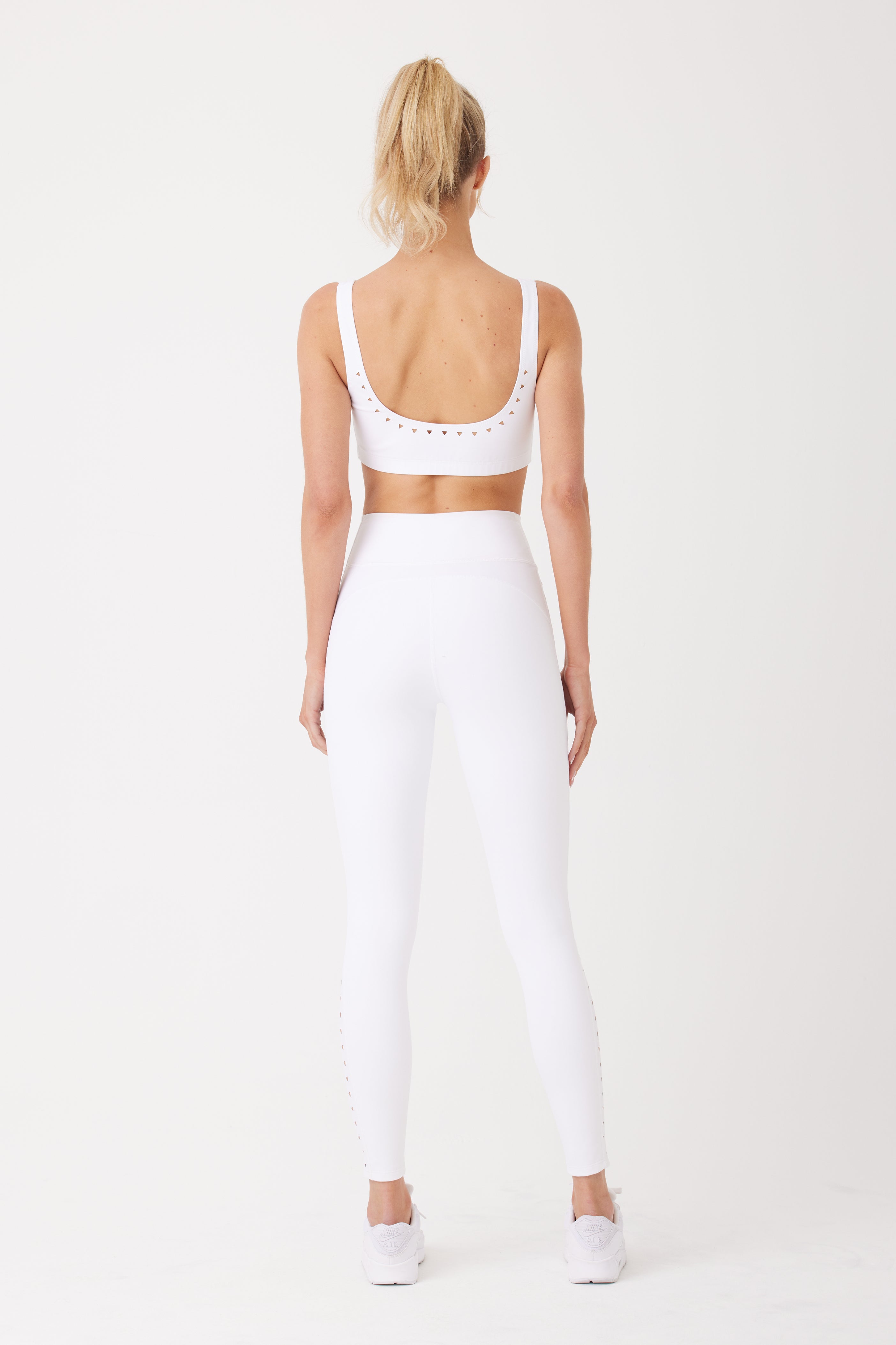 A person with a ponytail stands back to the camera in a white Emily Bra by Gigi C Sport, designed with breathable fabric and lasercut technology, featuring delicate edge cutouts. The ensemble is paired with sleek white sneakers against a plain white backdrop.