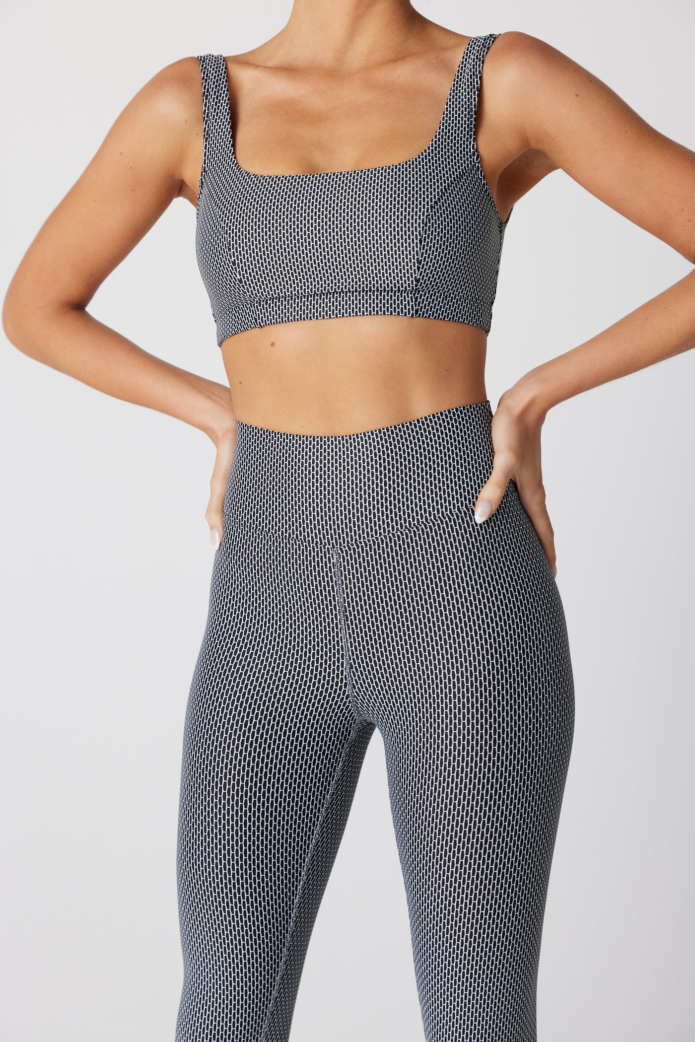 A person confidently poses hands on hips in a matching two-piece outfit featuring the Gigi C Sport Lori Legging, crafted from compression fabric with a textured black and white pattern. The high-waisted design highlights their form against a plain white background.