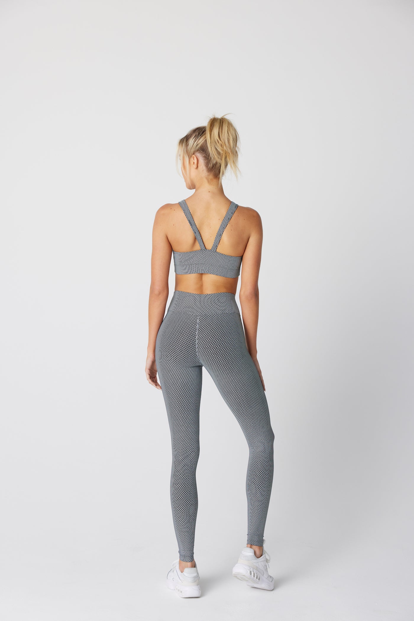 A person with a blonde ponytail faces away, wearing a grey Gigi C Sport Nicki Top and leggings made from Italian fabric with a honeycomb jacquard pattern, paired with white sneakers against a plain light background.