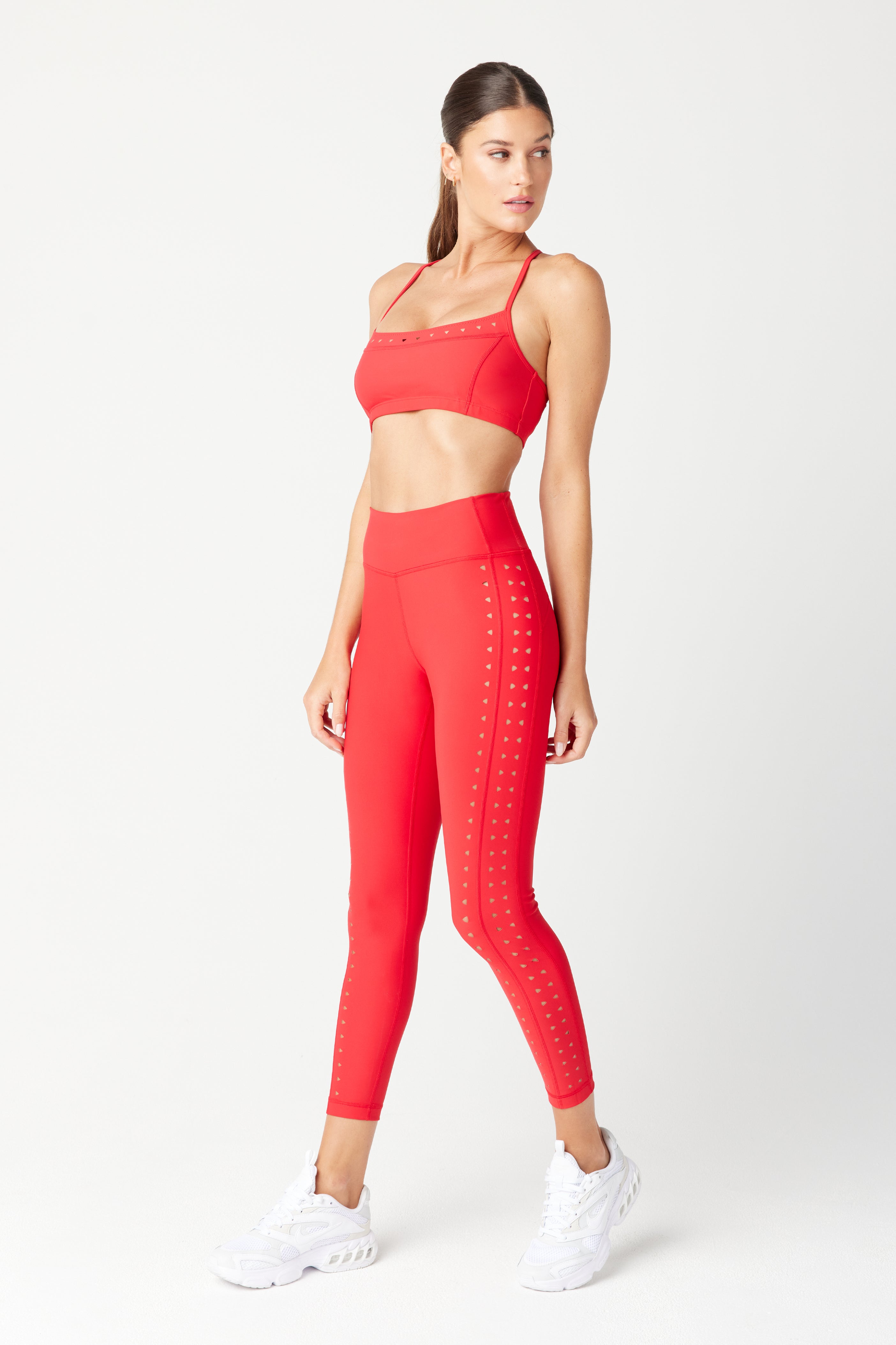 A woman confidently wears a red athletic outfit featuring the Giselle Bra by Gigi C Sport and high-waisted leggings. Made from breathable recycled fabric with cutout details, she completes the look with white athletic shoes against a plain white background.