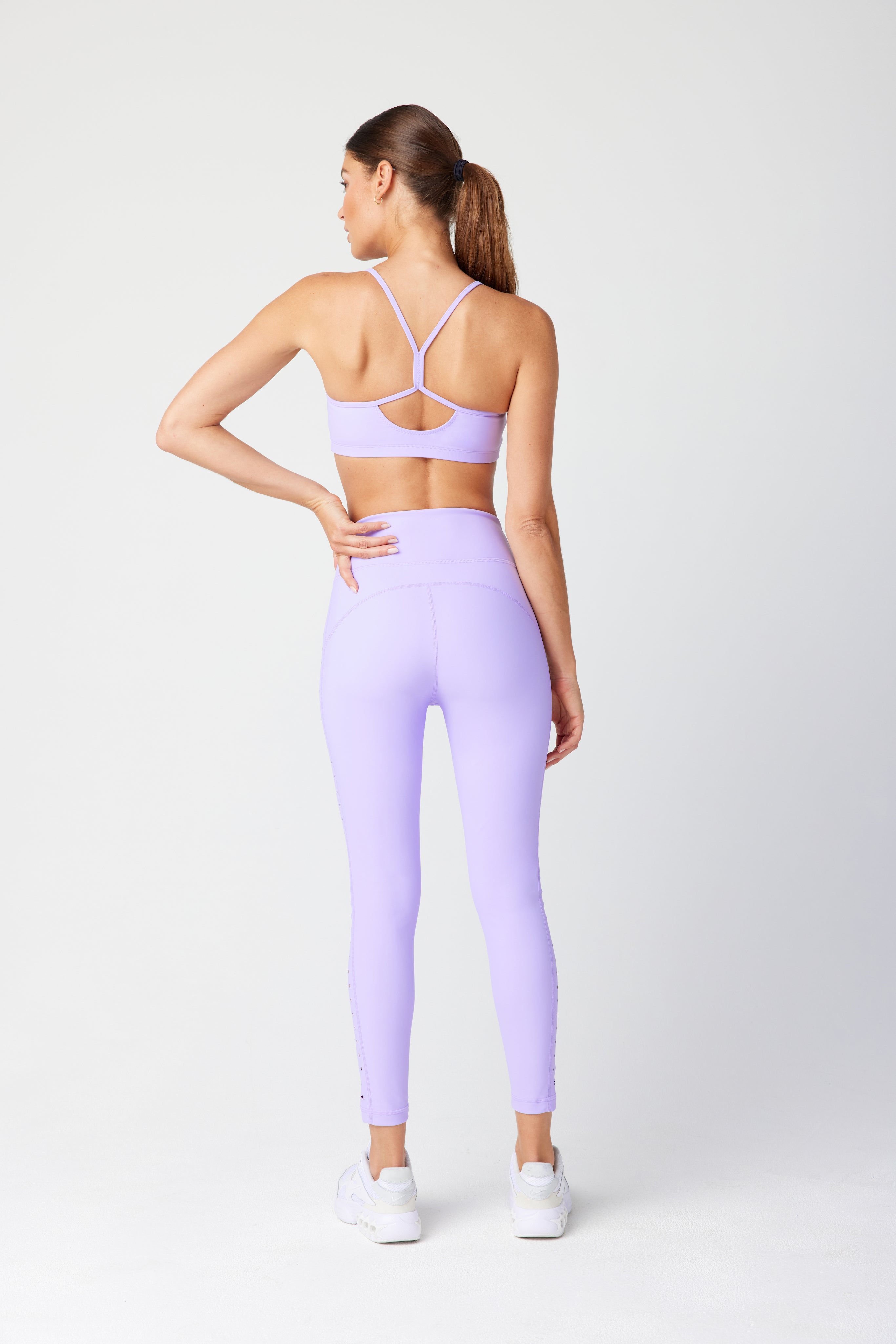 Facing away, a person wears Gigi C Sports Giselle Bra in lavender and leggings crafted from recycled fabric, complemented by white sneakers. Their brown hair is styled in a ponytail against a simple light background.