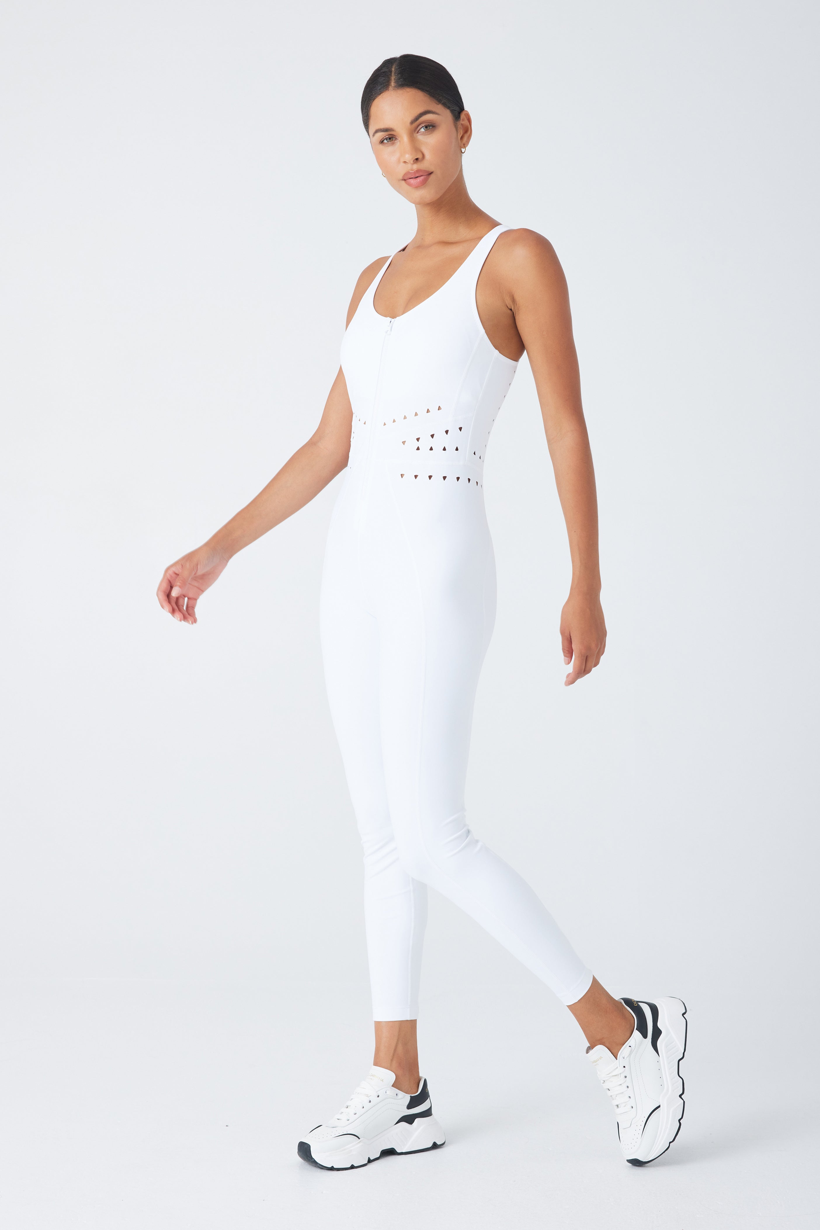 A person stands confidently against a plain background, wearing the form-fitting Hunter Jumpsuit by Gigi C Sport, featuring cutout design details, paired with white sneakers. Their hair is pulled back, and one arm rests relaxed by their side.