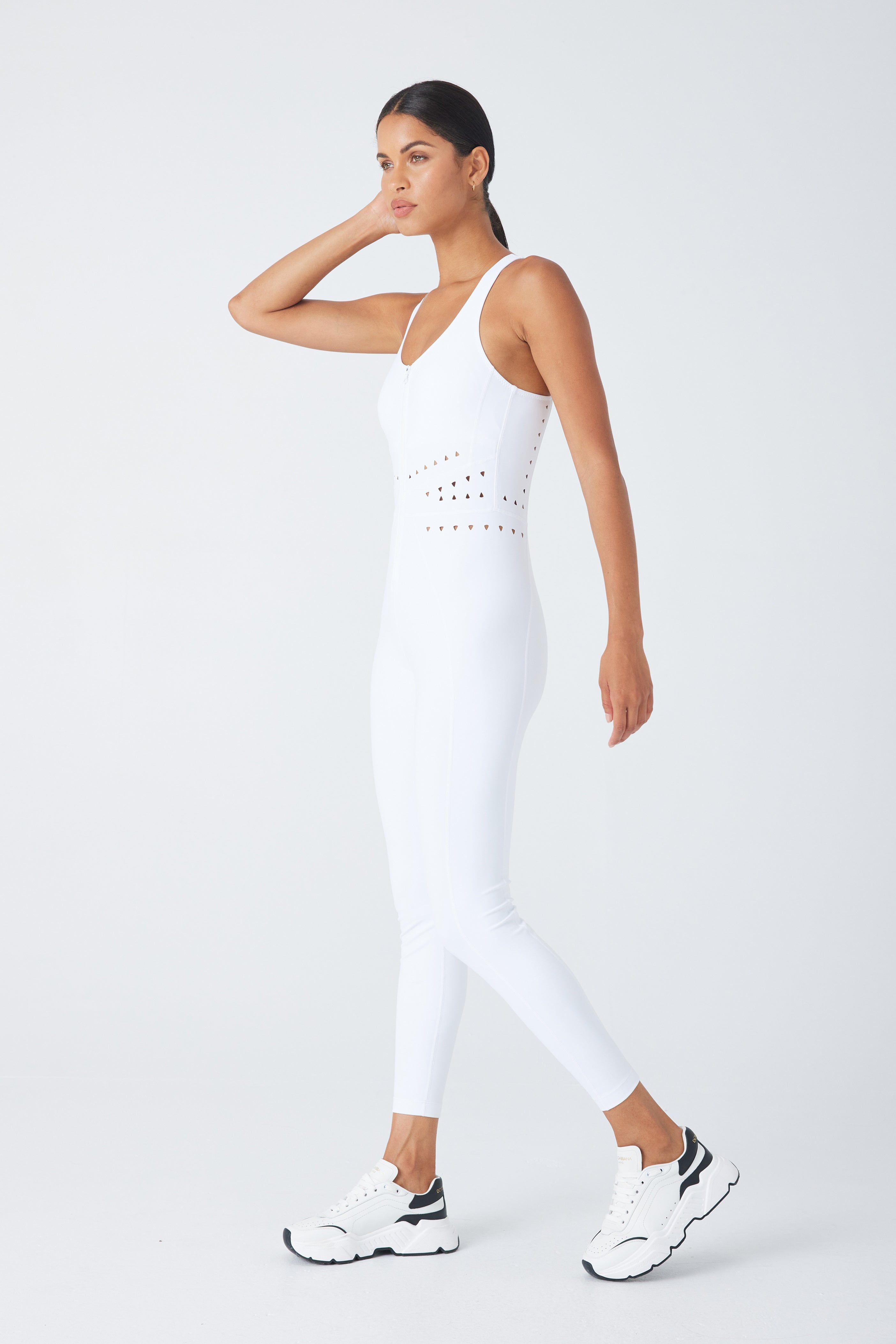 A person models the sleek Hunter Jumpsuit by Gigi C Sport, featuring a stylish cutout design. Theyre paired with black and white sneakers, striking an active pose while touching their head against a plain studio backdrop.