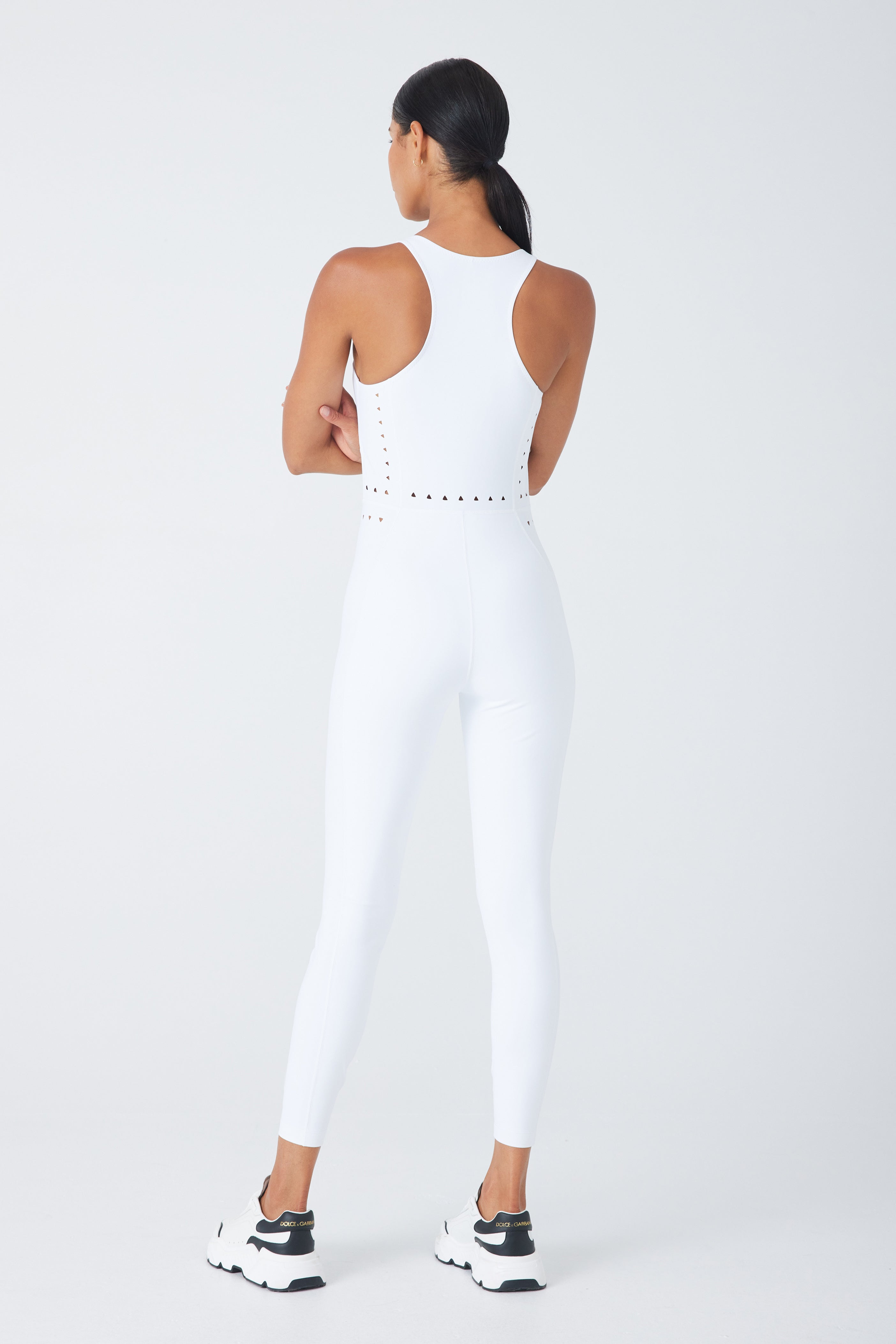 A woman, back to the camera against a crisp white backdrop, wears the Gigi C Sport Hunter Jumpsuit, a white activewear piece with a sleek cutout design. She pairs it with black-and-white sneakers and has her arms crossed and dark hair tied back.
