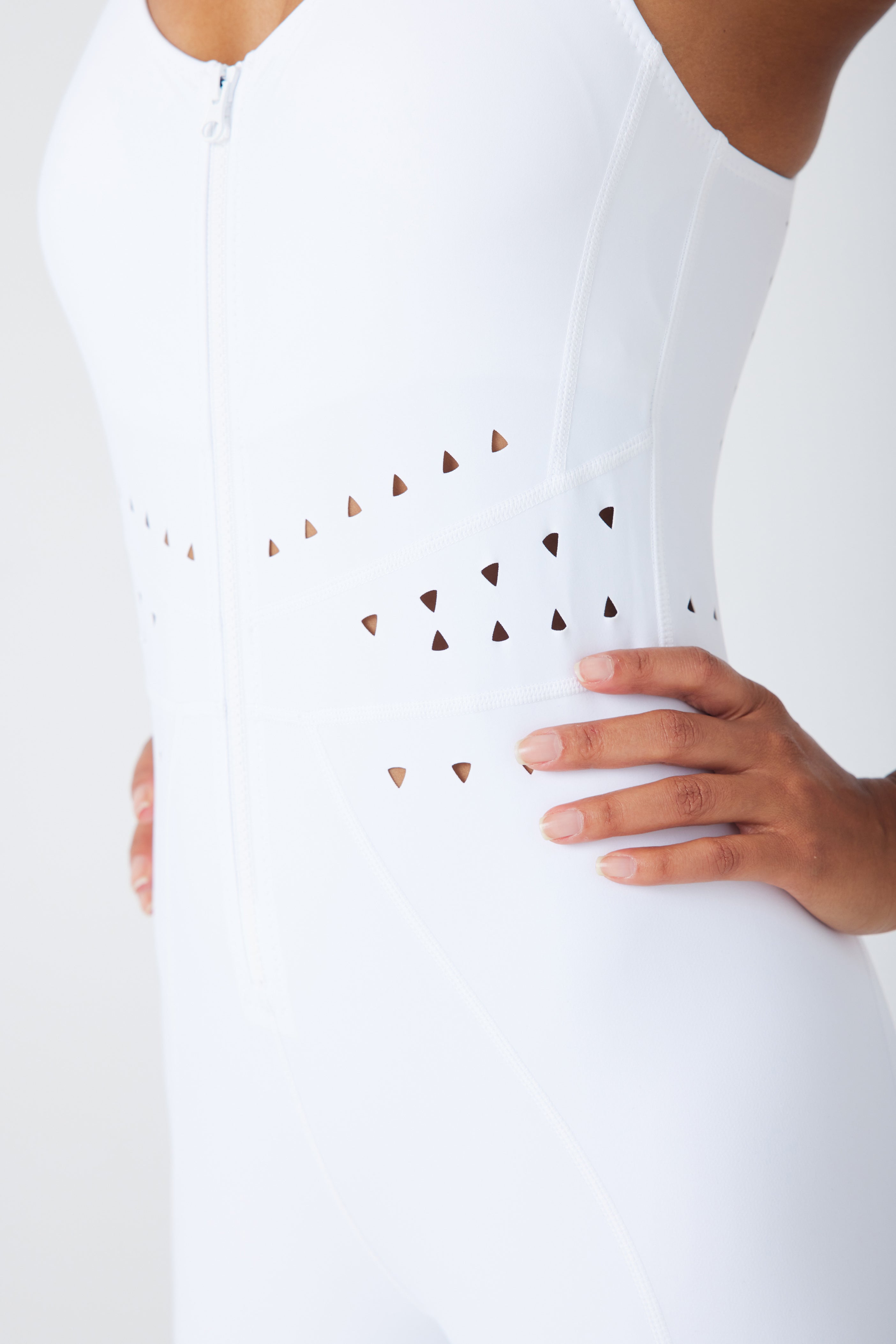 A close-up shows a person wearing the Hunter Jumpsuit by Gigi C Sport. This white sleeveless activewear features a front zipper and waist cutouts for added flair, with hands on hips highlighting its texture and fit.