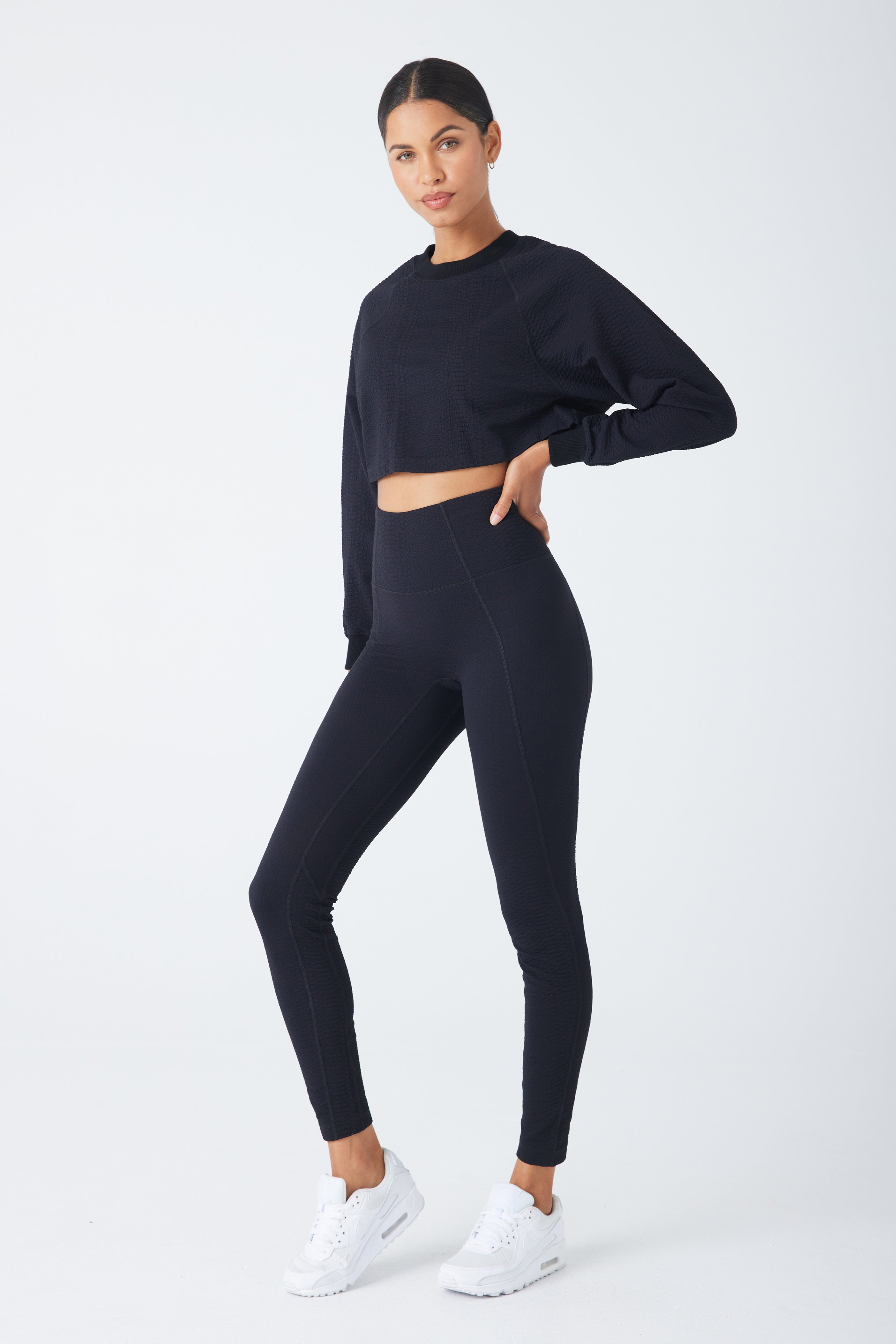 A woman confidently poses in a black long-sleeve crop top with high-waisted Ava Legging by Gigi C Sport, crafted from bi-stretch compression waffle jacquard fabric. Wearing white sneakers, she has her hand on her hip against a plain white backdrop.