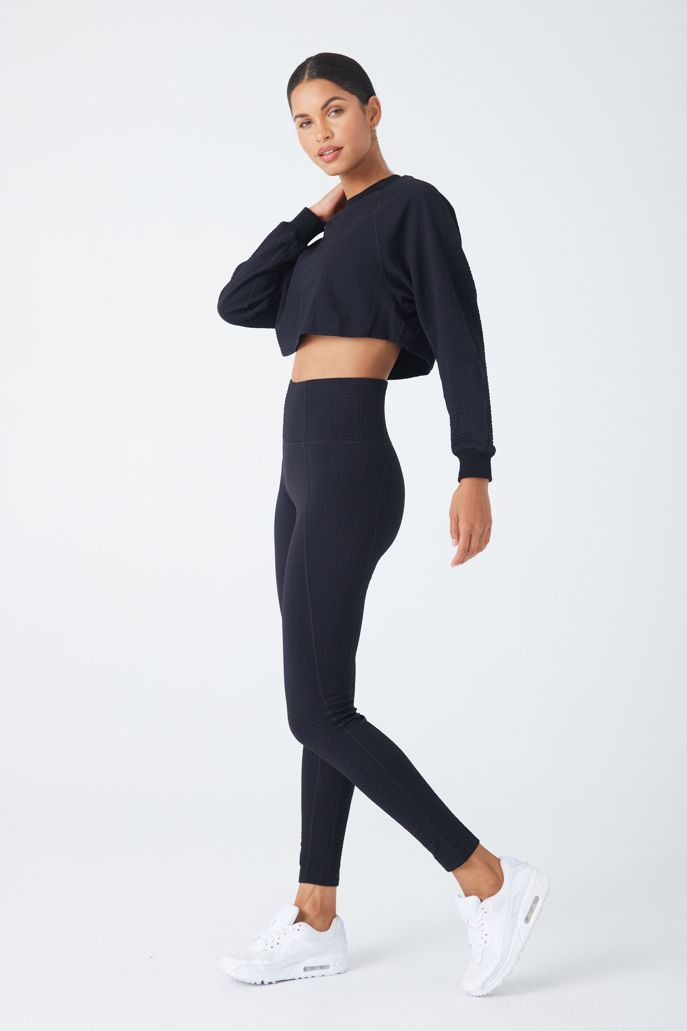 A person poses confidently against a plain white background, wearing a black crop top and Gigi C Sports high-waisted Ava Legging in bi-stretch compression waffle jacquard. White sneakers complete the look as they stand with one leg forward, gazing relaxedly at the camera.