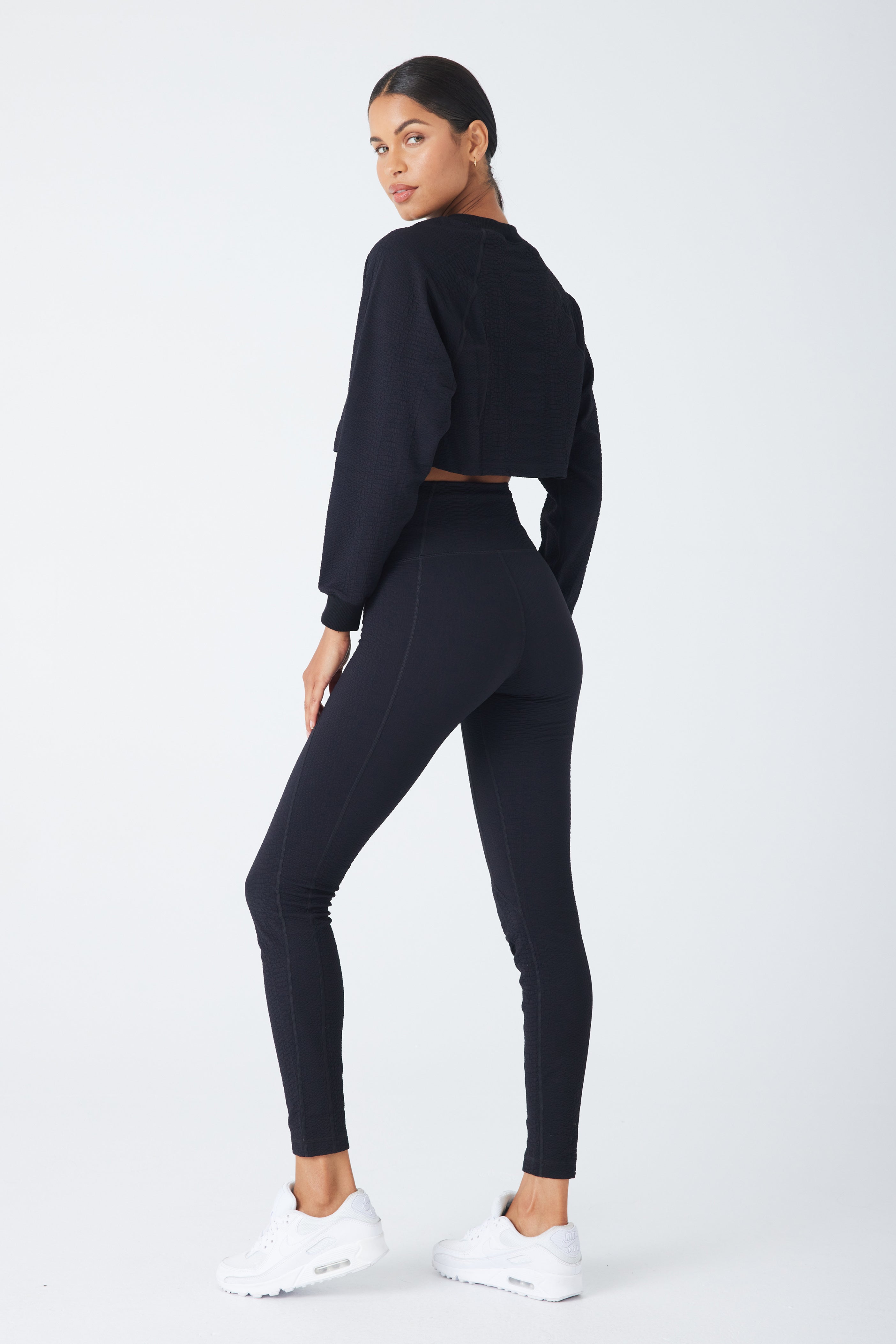 A person stands, glancing back in a Gigi C Sports Ava Legging set, showcasing a black long-sleeve cropped top and matching high-waisted leggings made from bi-stretch compression waffle jacquard fabric. The look is finished with white sneakers against a plain light background.
