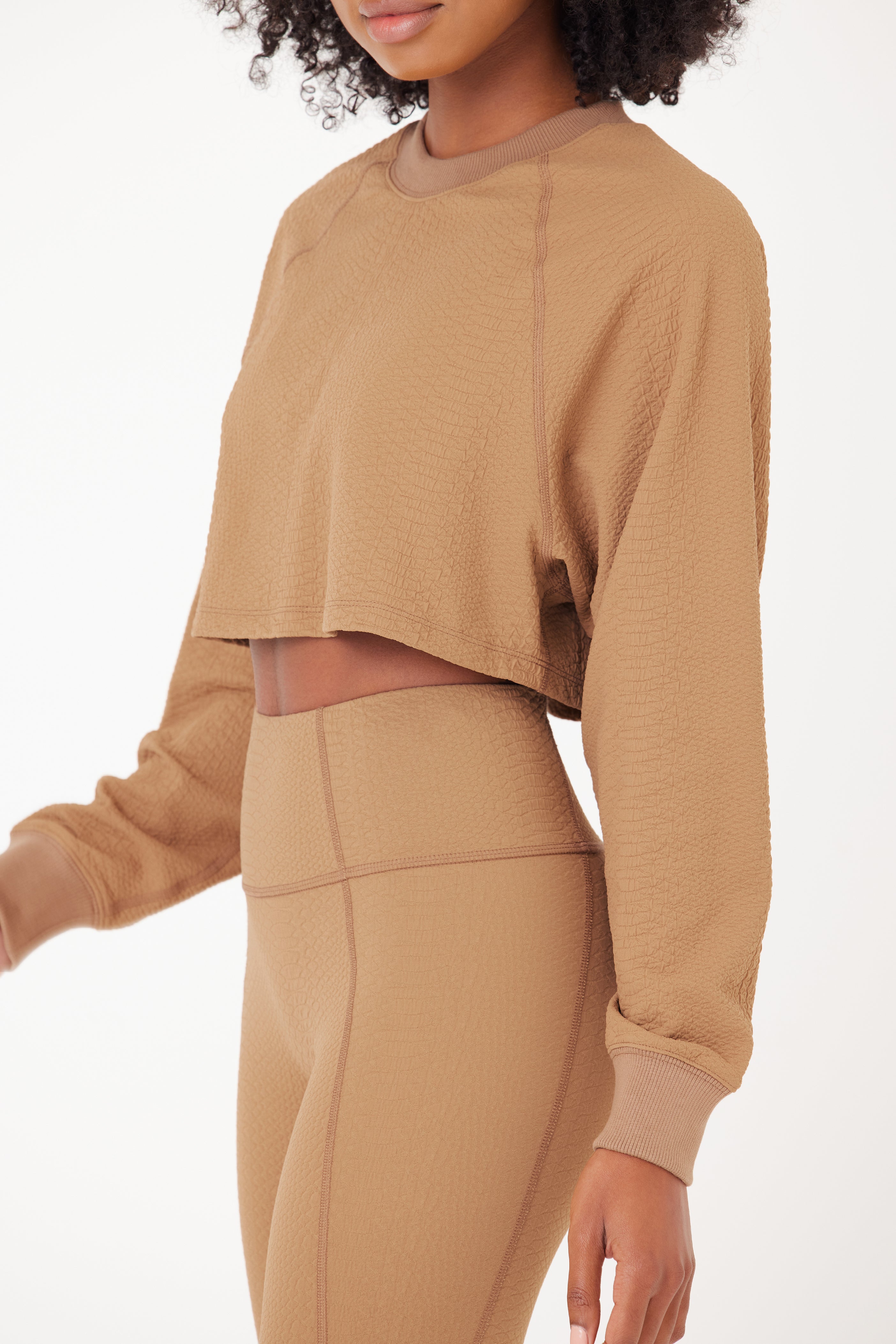 A woman models a light brown textured, long-sleeve cropped top and high-waisted leggings, her natural curls cascading as she exudes effortless style. Her look is reminiscent of the Isabella Pullover by Gigi C Sport, adding flair to her workout ensemble against a plain white background.