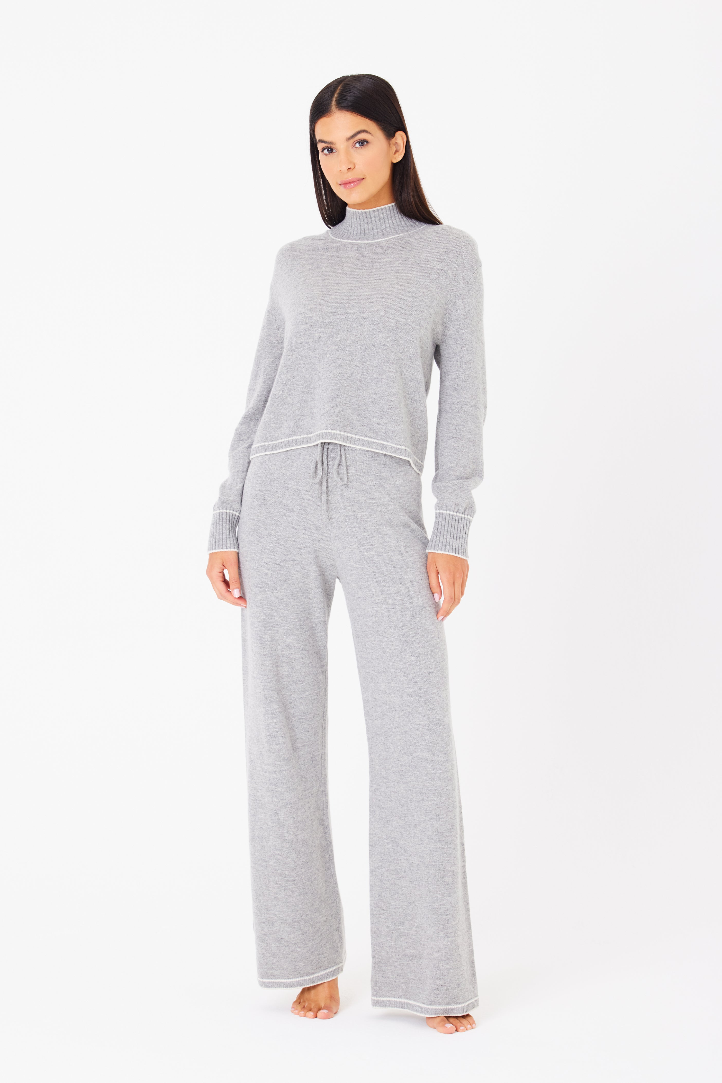 A person with long, dark hair stands barefoot on a white background in a Gigi C Lounge set. The light gray knit sweater and Aubrie Wide Leg Pant, made from a wool-cashmere blend, showcase a loose fit with ribbed cuffs and drawstring waist.