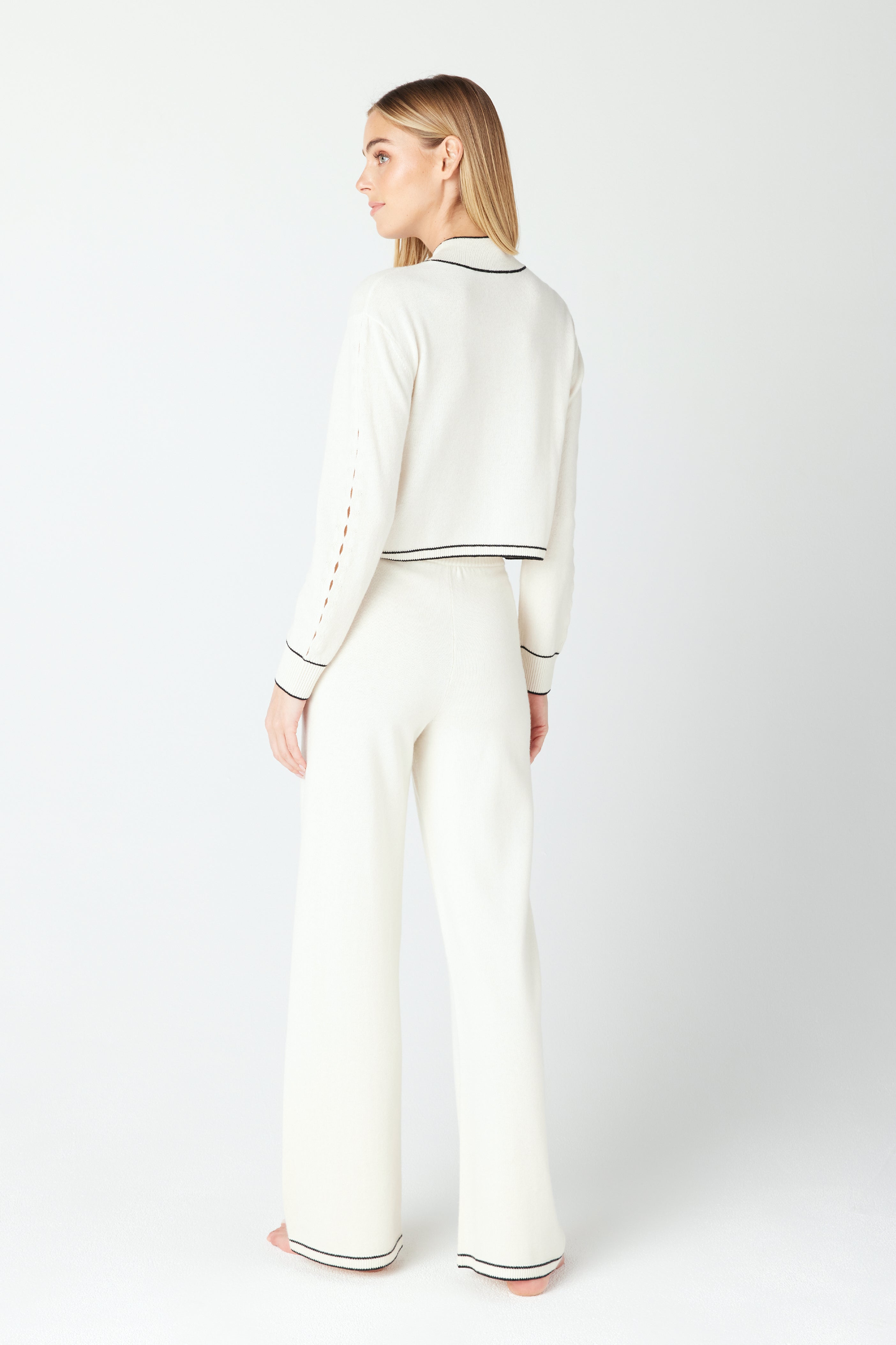 A woman with long blonde hair stands barefoot on a light background, facing away, wearing the Jacqueline Pullover by Gigi C Lounge, crafted from recycled cashmere. The cream-colored set features black trim and teardrop cut-out details on the wide-legged pants and matching top.