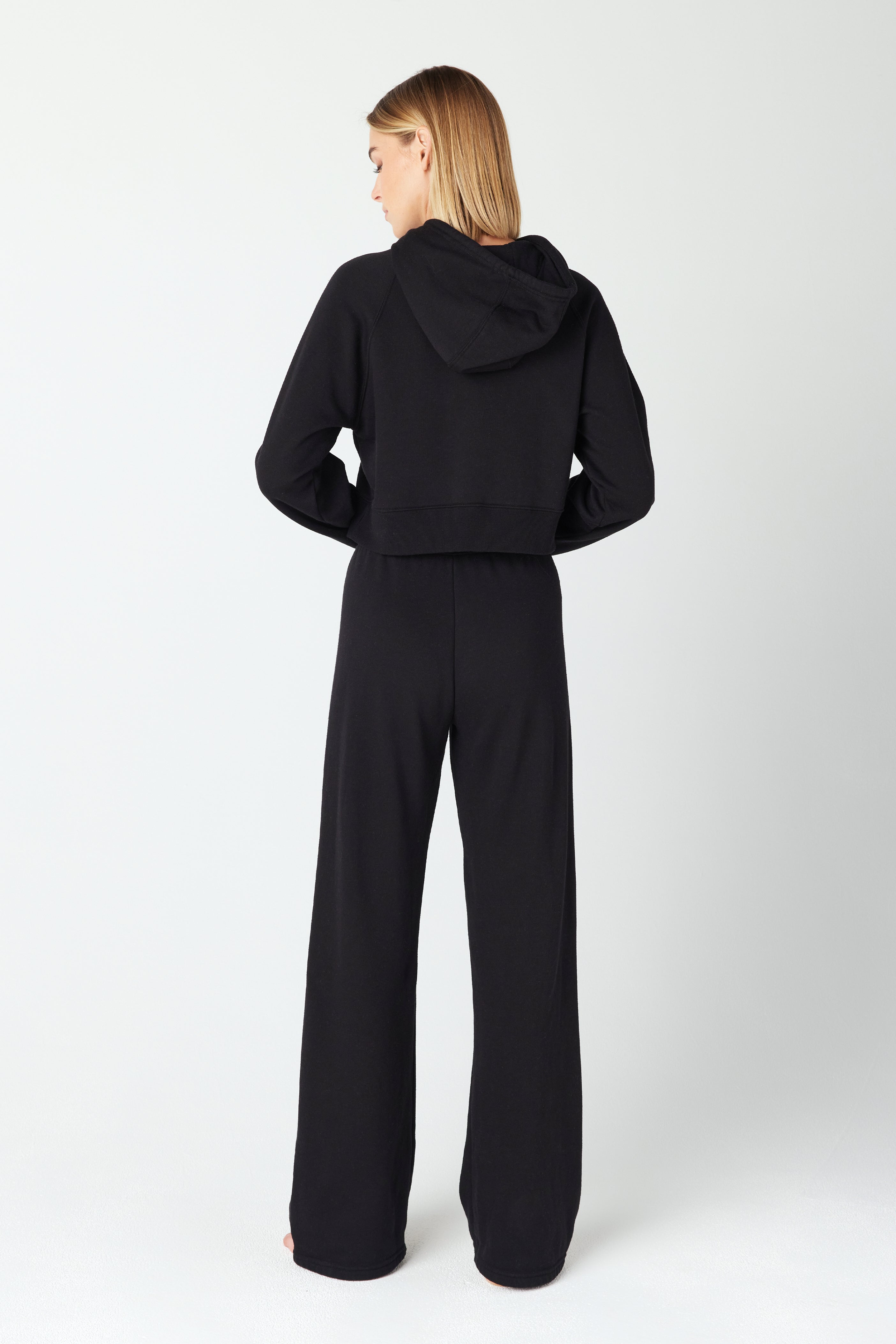 A person faces away from the camera, dressed in a black Juliet hoodie and wide-leg Harlow Pant by Gigi C Lounge against a plain light gray background.