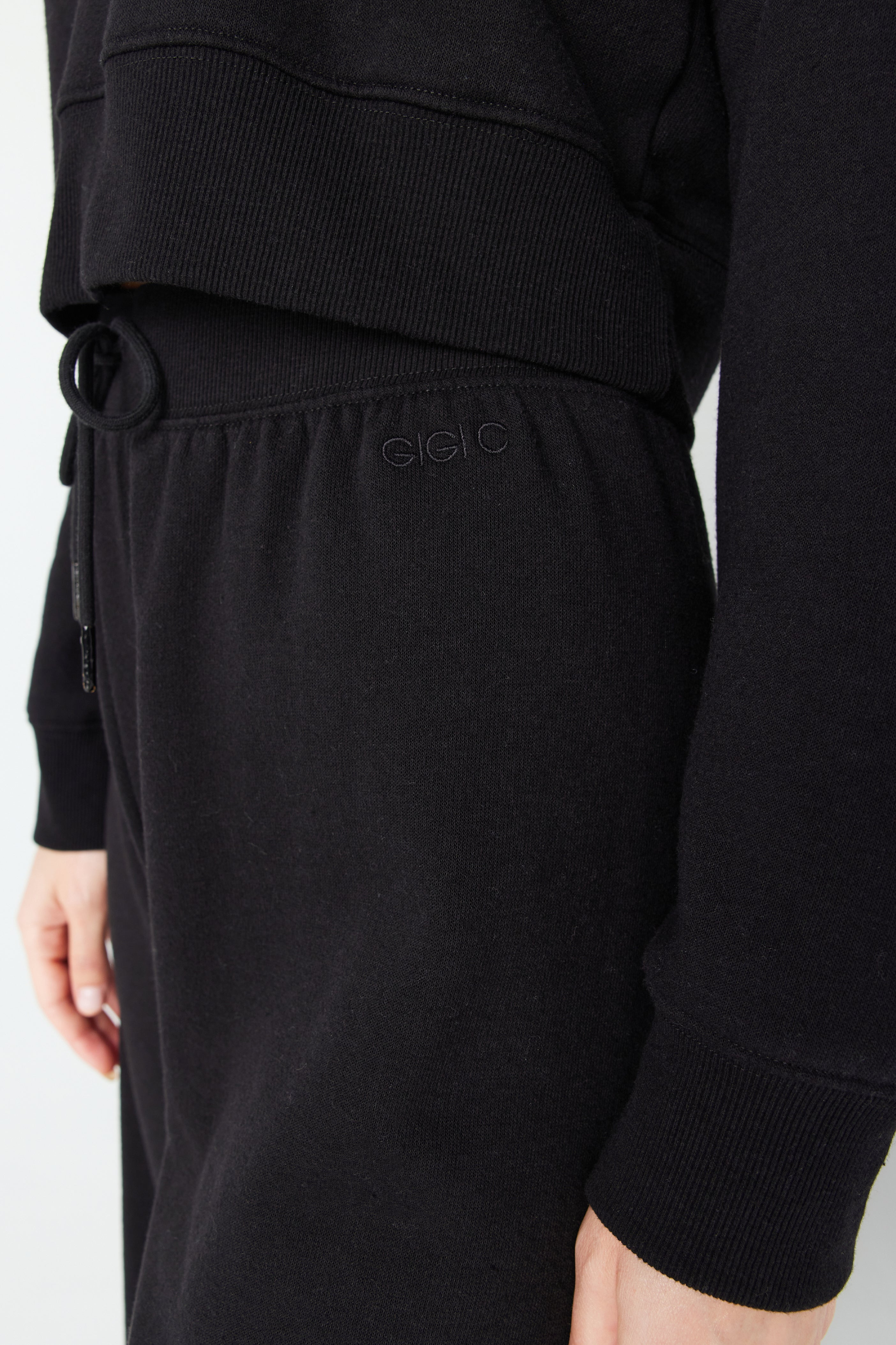 A person is seen from the side and partial upper body, wearing a black Kenzie pullover paired with Gigi C Lounges Harlow Pant, both featuring a subtle embossed logo. The outfit exhibits a relaxed, casual fit.