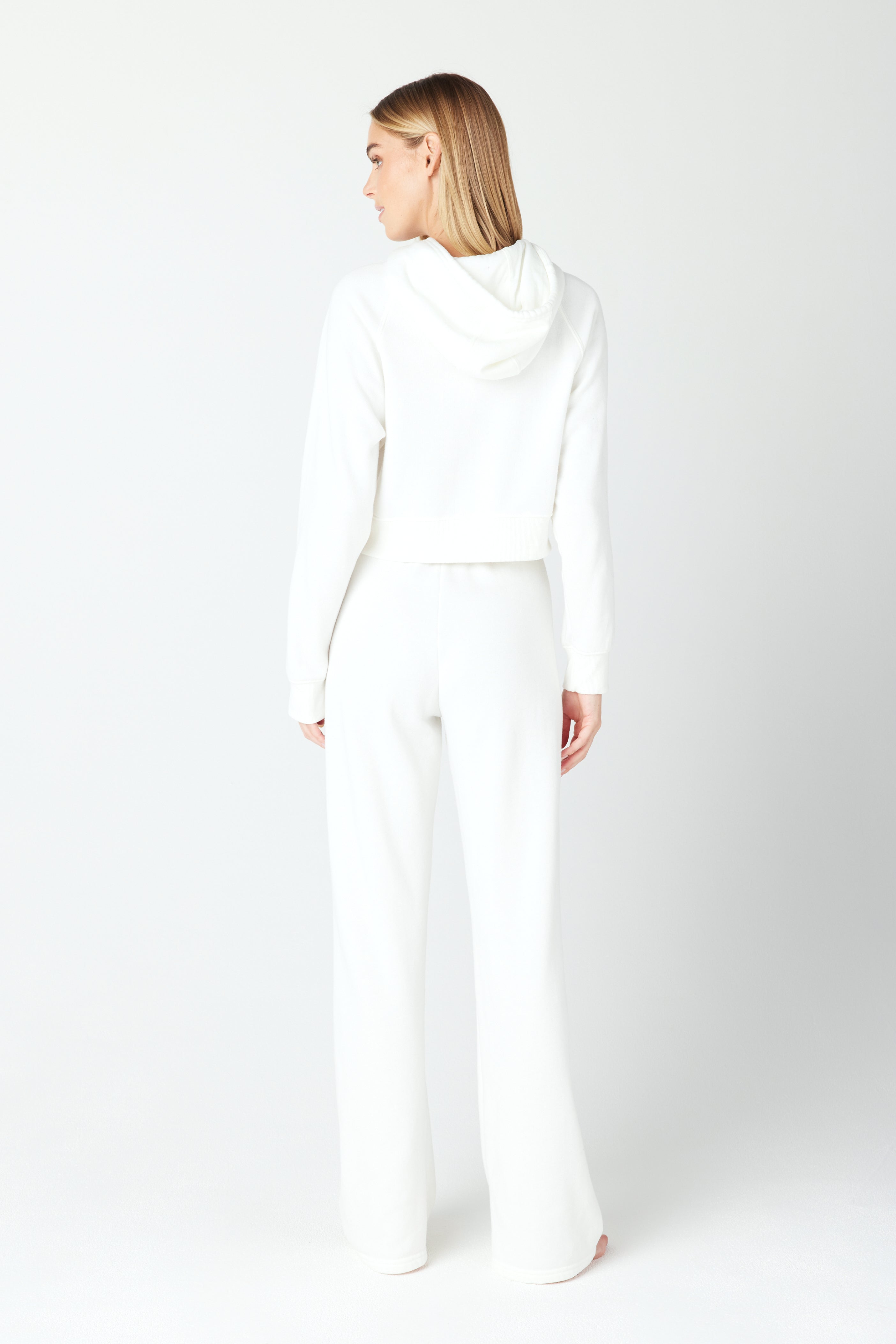 A person in a white Juliet hoodie and wide-leg Gigi C Lounge Harlow Pant stands facing away, glancing left. The plain white backdrop complements the casual and comfortable style of their outfit.