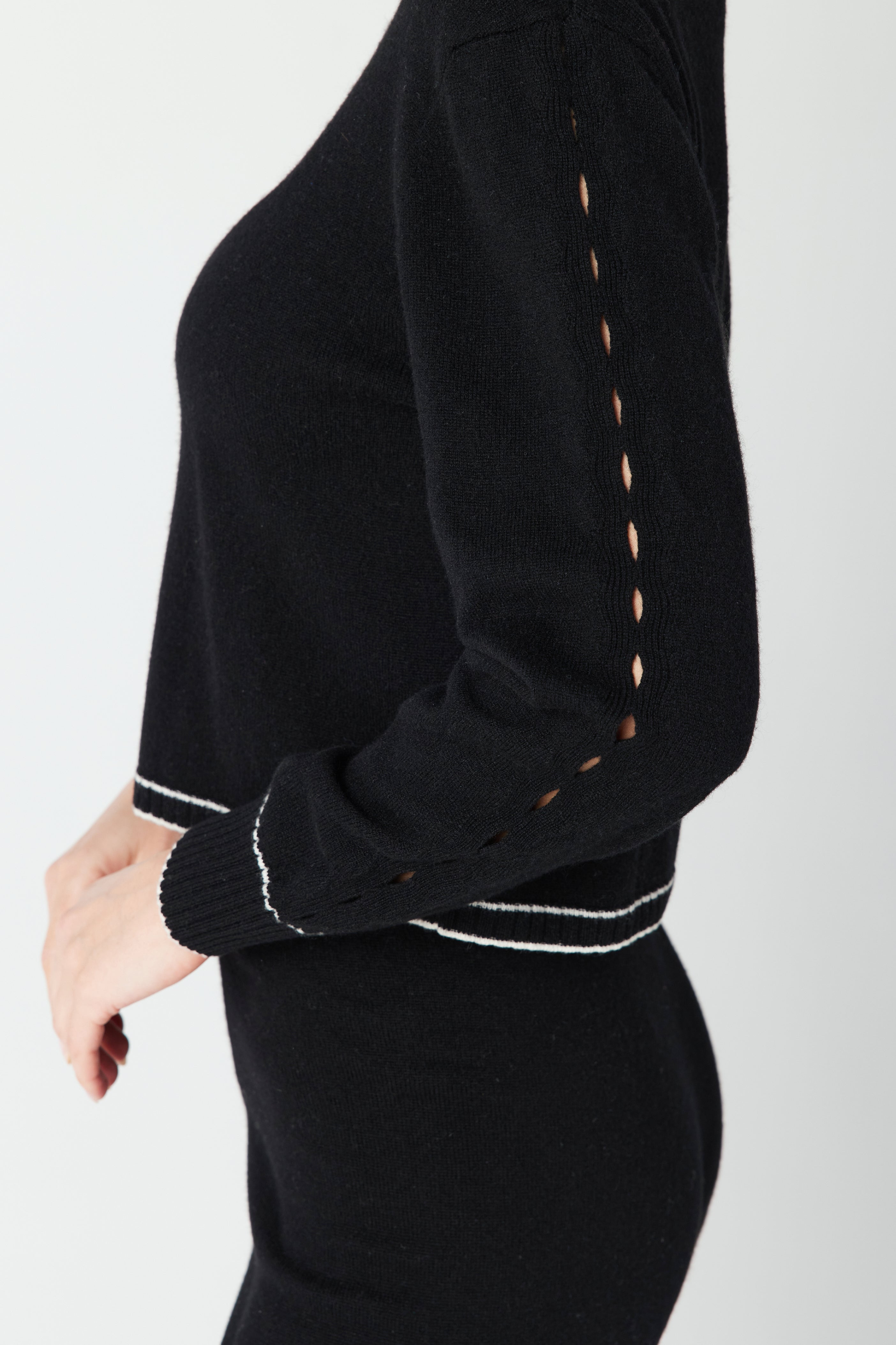 A person wearing the Gigi C Lounge June Sweater and a matching skirt in a luxurious wool and recycled cashmere blend showcases openwork detailing on the sleeves, with white trim at cuffs and waist. The ribbed knit hem is highlighted as they stand in profile, hand on hip.