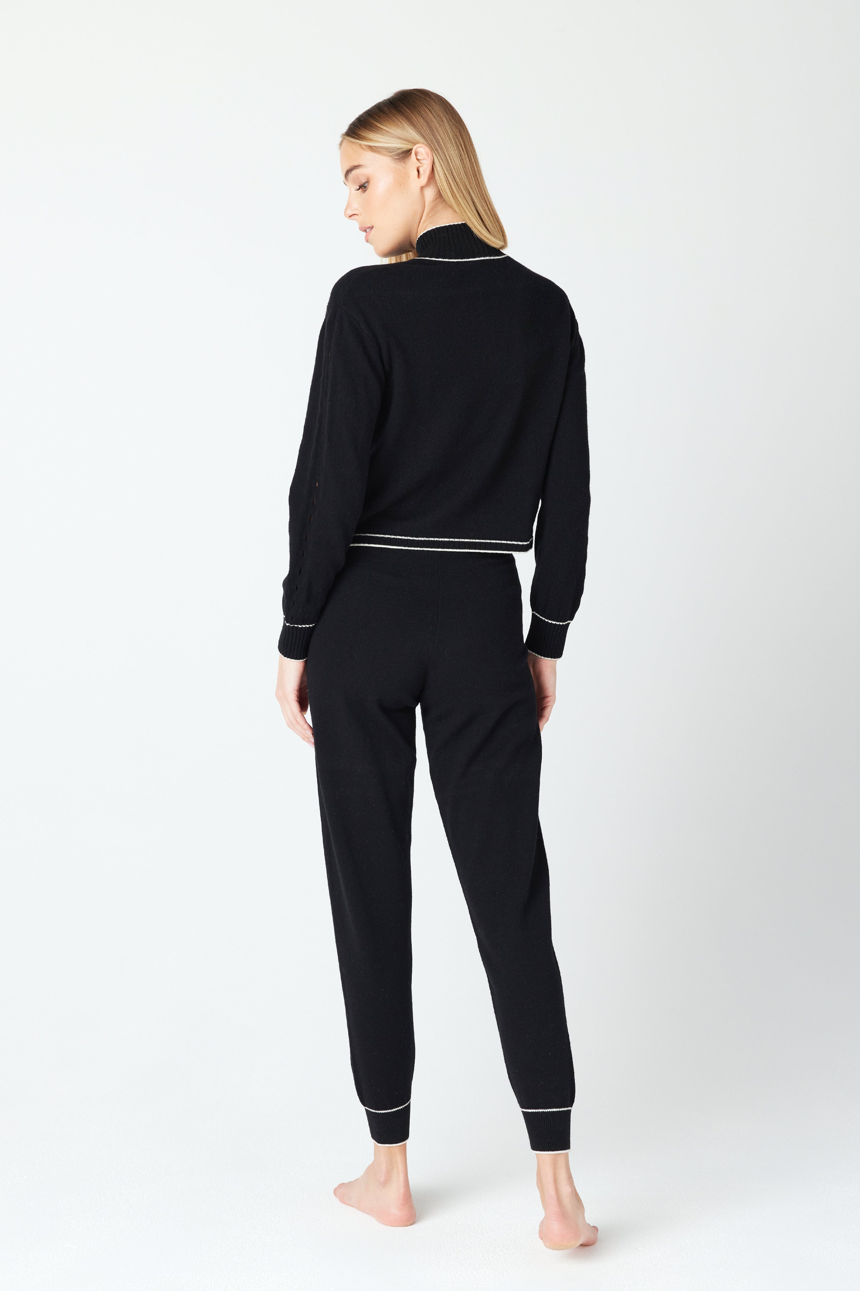 Standing barefoot and facing away from the camera, a person with long hair showcases the chic Gigi C Lounge June Sweater in black, featuring white trim and a ribbed knit hem. Crafted from a luxurious wool and recycled cashmere blend, the plain white background enhances its elegance.