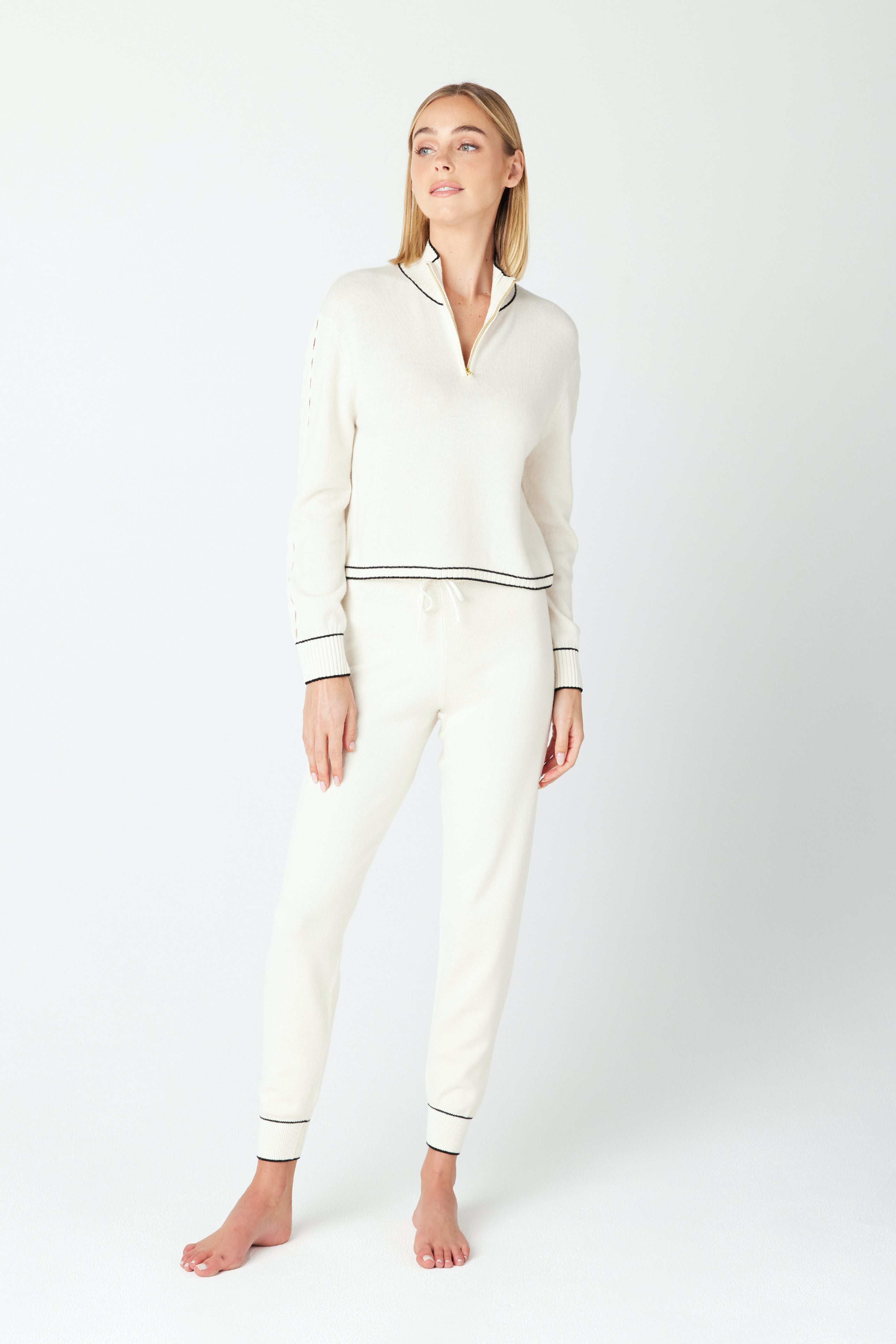 A person stands against a plain white background, wearing the stylish June Sweater set by Gigi C Lounge—a sleek white loungewear outfit with black trim. It includes a cozy long-sleeve top with a small collar and matching pants. The barefoot individual has long hair.