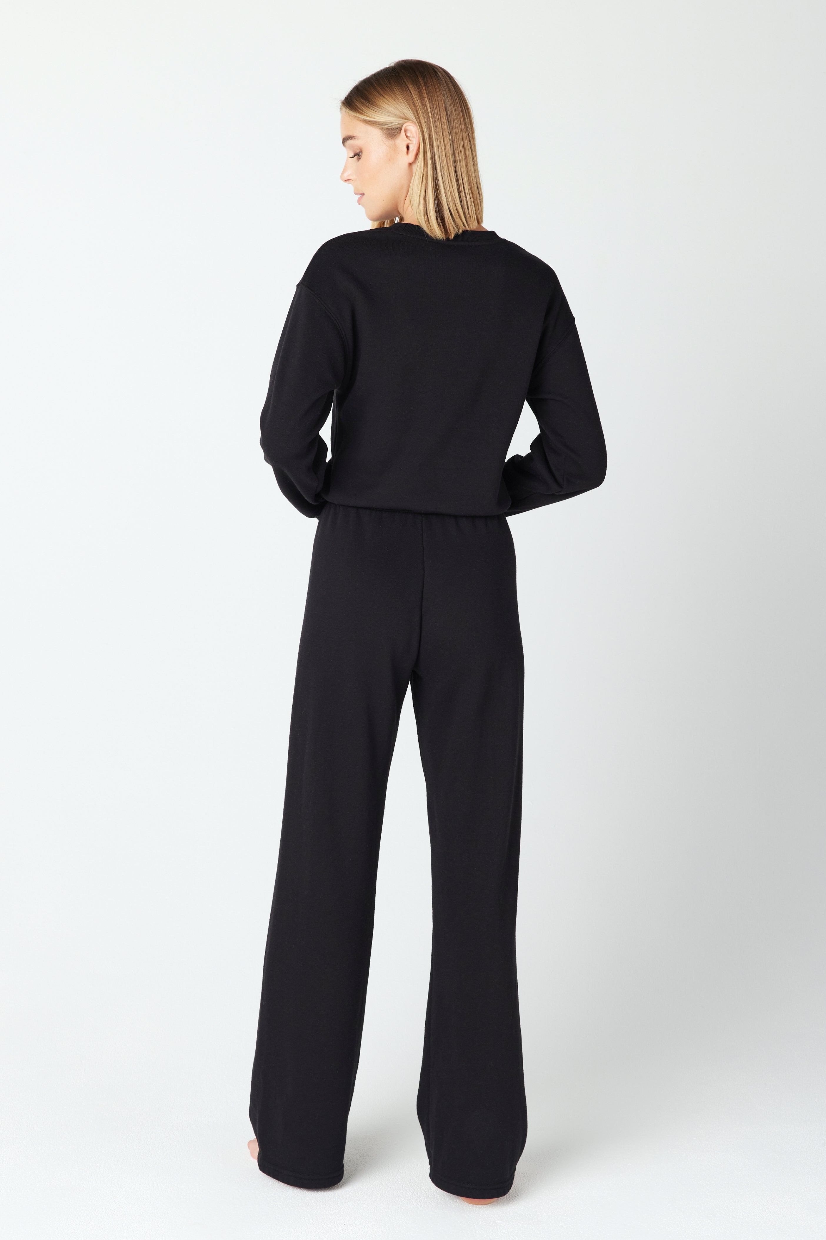 A person with straight, medium-length blonde hair faces away from the camera against a plain white background, wearing a stylish black Kenzie Pullover from Gigi C Lounge paired with matching black pants.