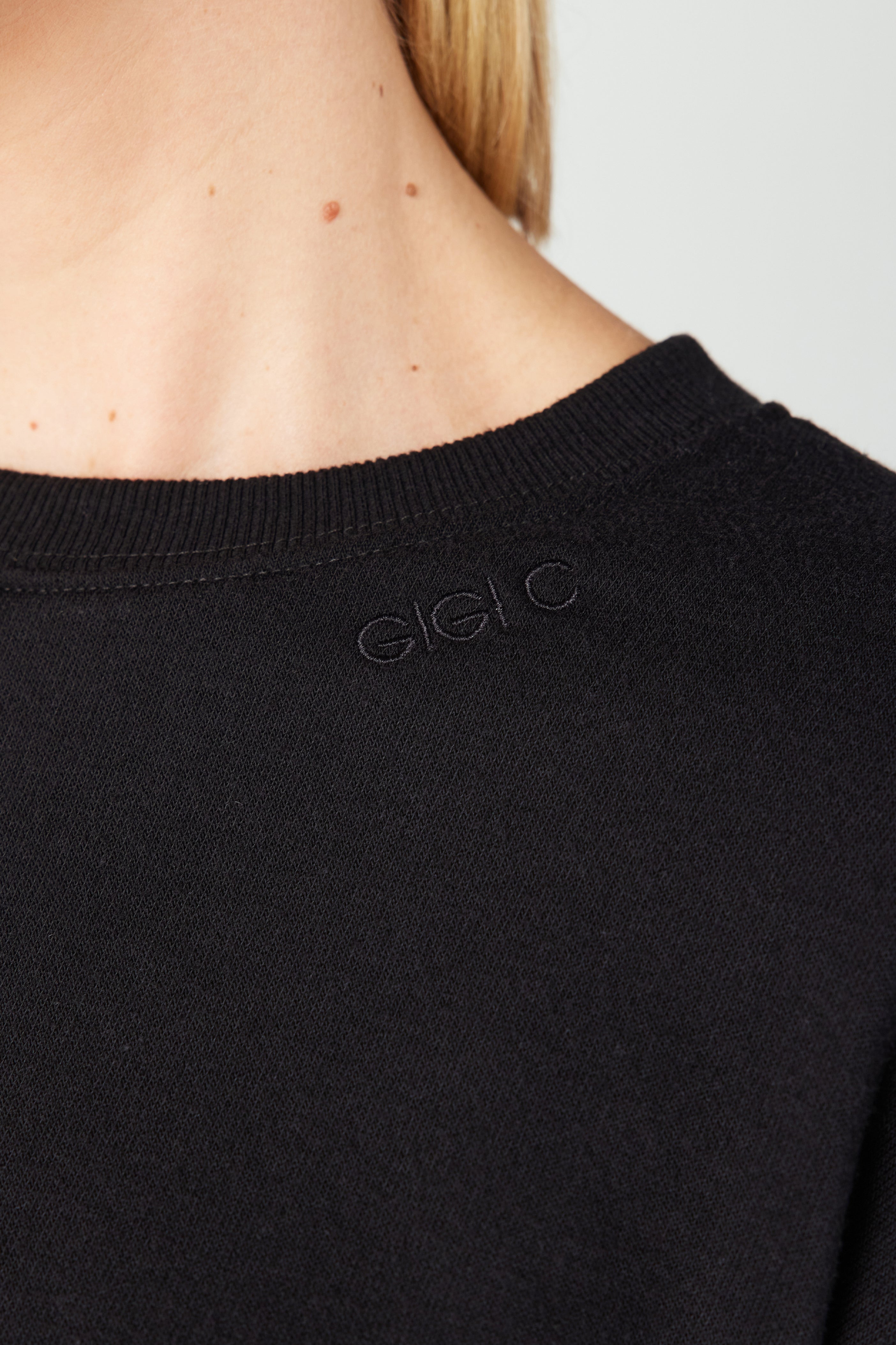 A close-up of the Kenzie Pullover by Gigi C Lounge shows Gigi C embroidered in subtle black thread on a black garment, partially revealing a persons light skin neck and shoulder with moles.
