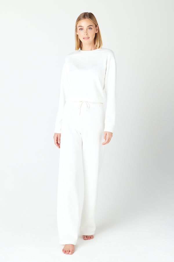 Against a plain backdrop, a barefoot individual with medium-length hair wears Gigi C Lounges white Kenzie Pullover and matching wide-leg pants in Loop Terry Fleece, creating a casual and relaxed style ideal for leisurely moments.