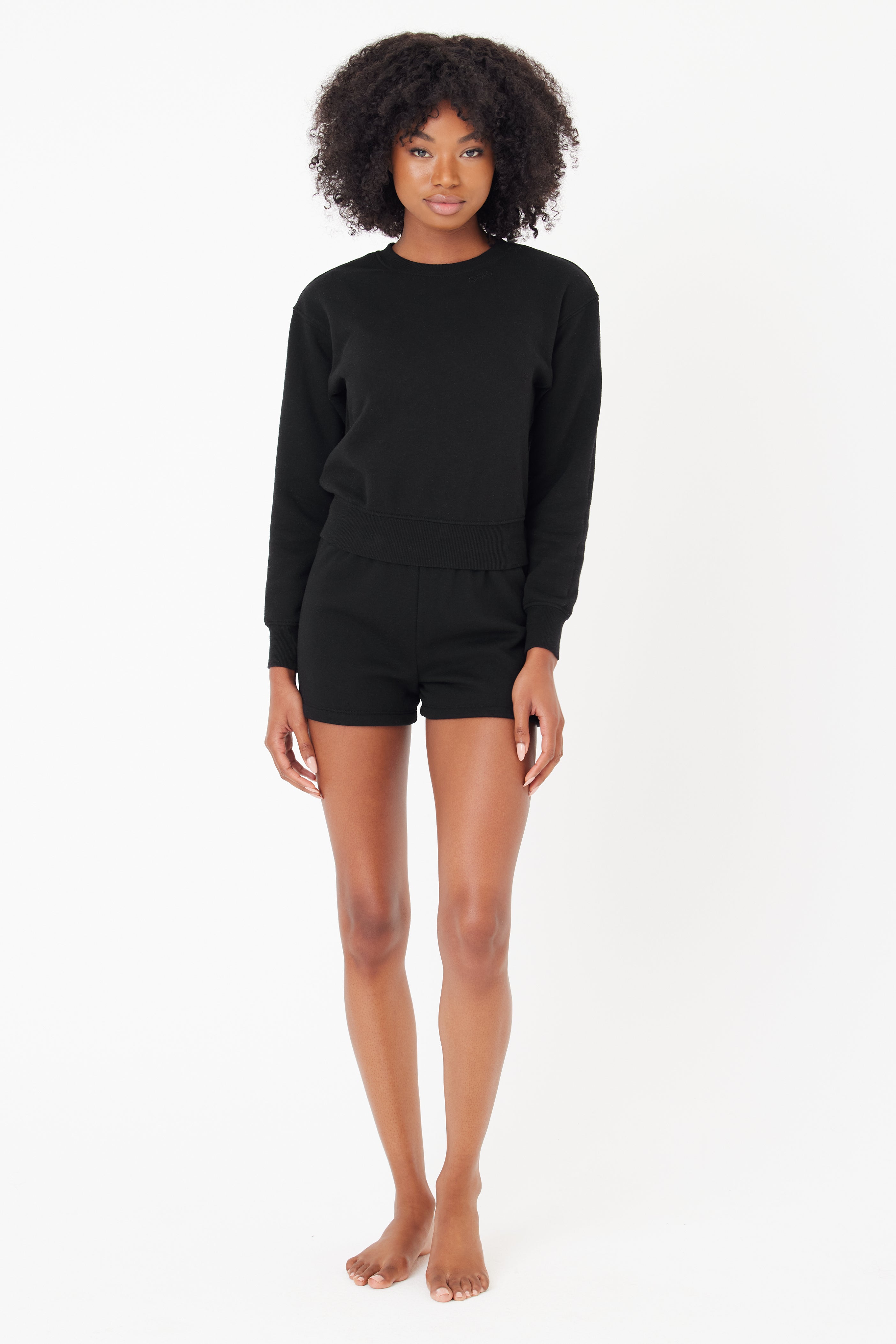 Someone with curly hair is wearing the Gigi C Lounge Louise Short and a black long-sleeve top from the new loungewear collection, standing barefoot against a plain white background.