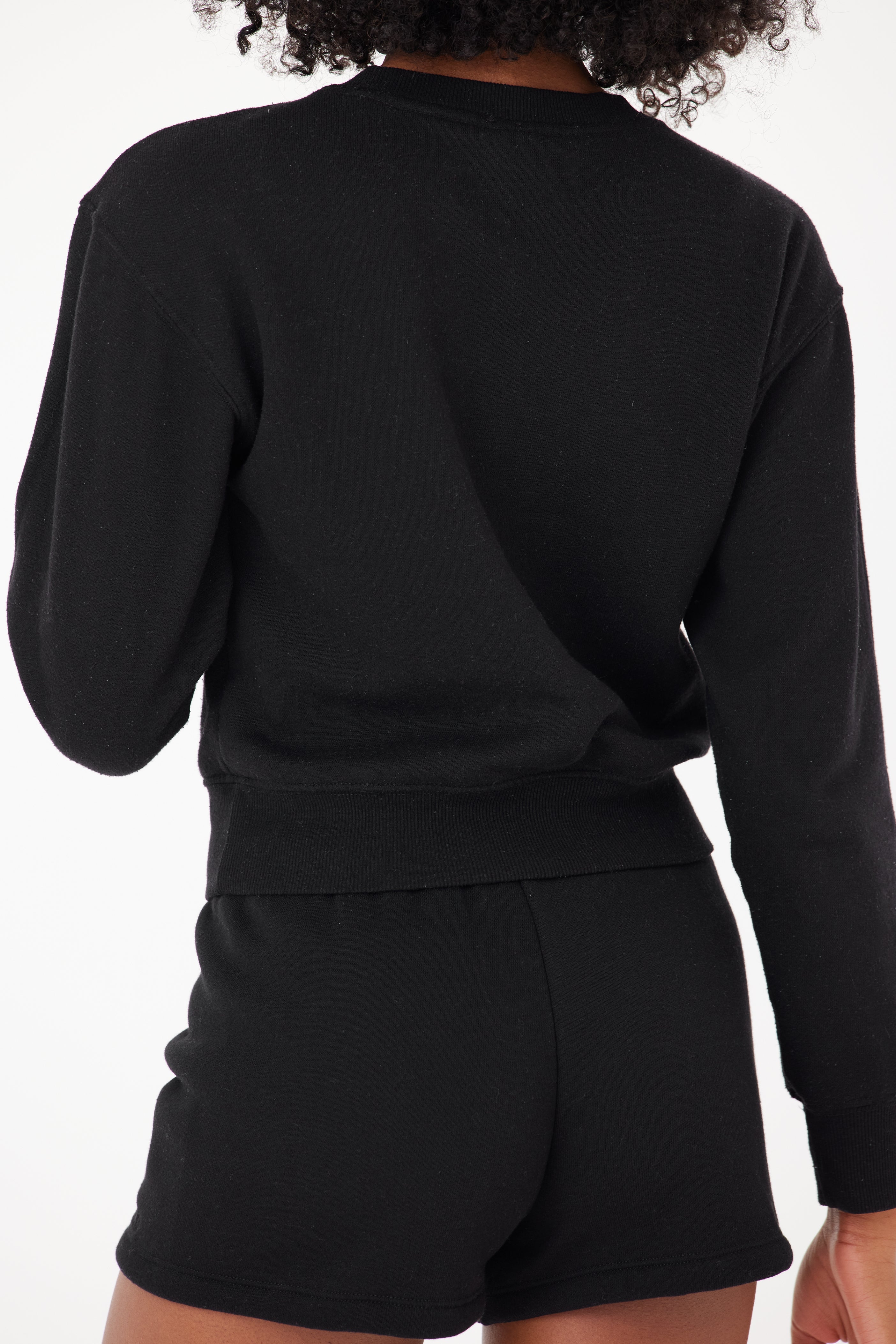 A person with curly hair, wearing a black long-sleeve top and the Louise Short by Gigi C Lounge, faces away from the camera against a plain white background.