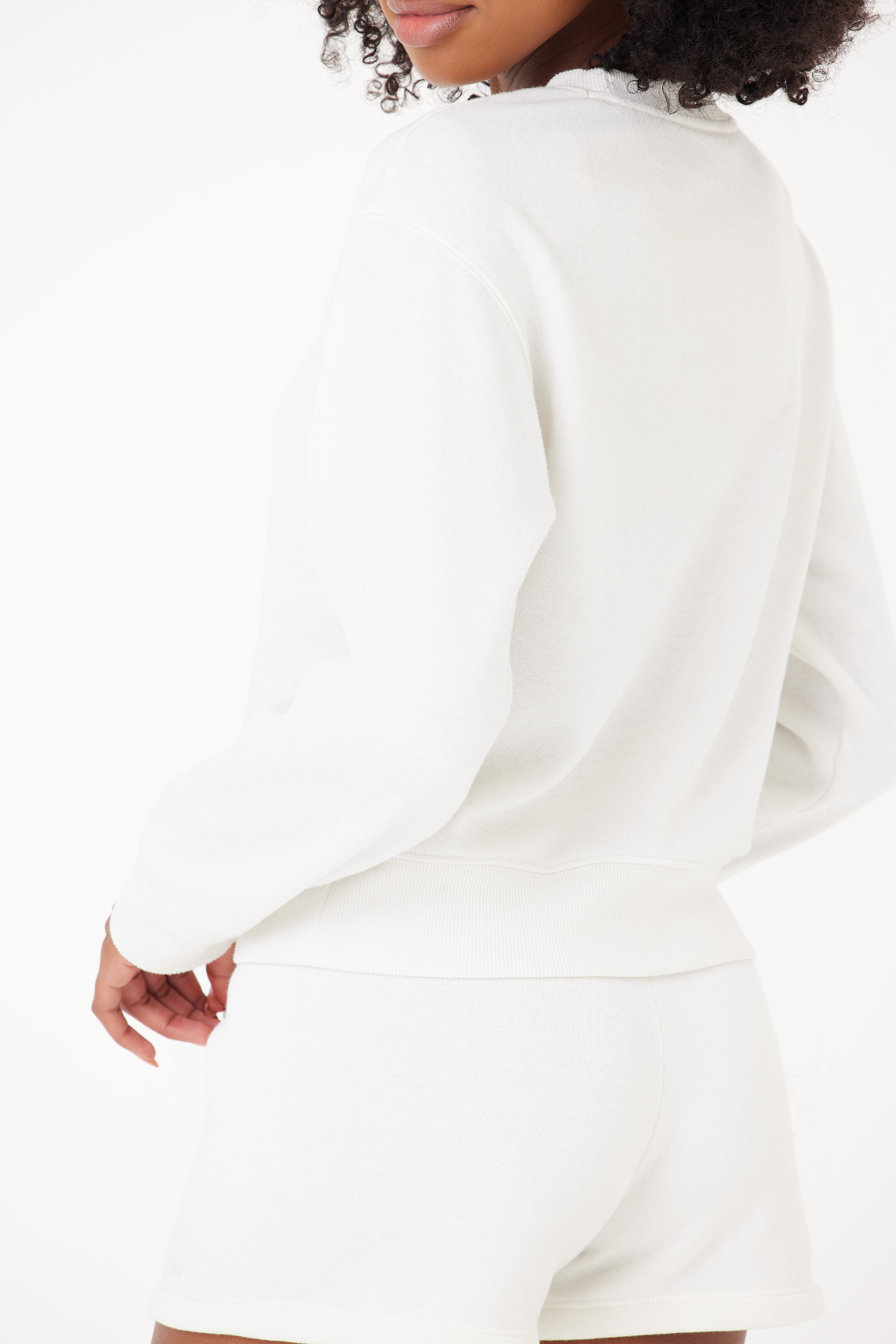 Against a white background, a person with curly hair partially visible is showcased in the Louise Short by Gigi C Lounge, paired with a white sweatshirt. The side view highlights the embroidered logo on their comfortable outfit, adding a subtle touch of style.