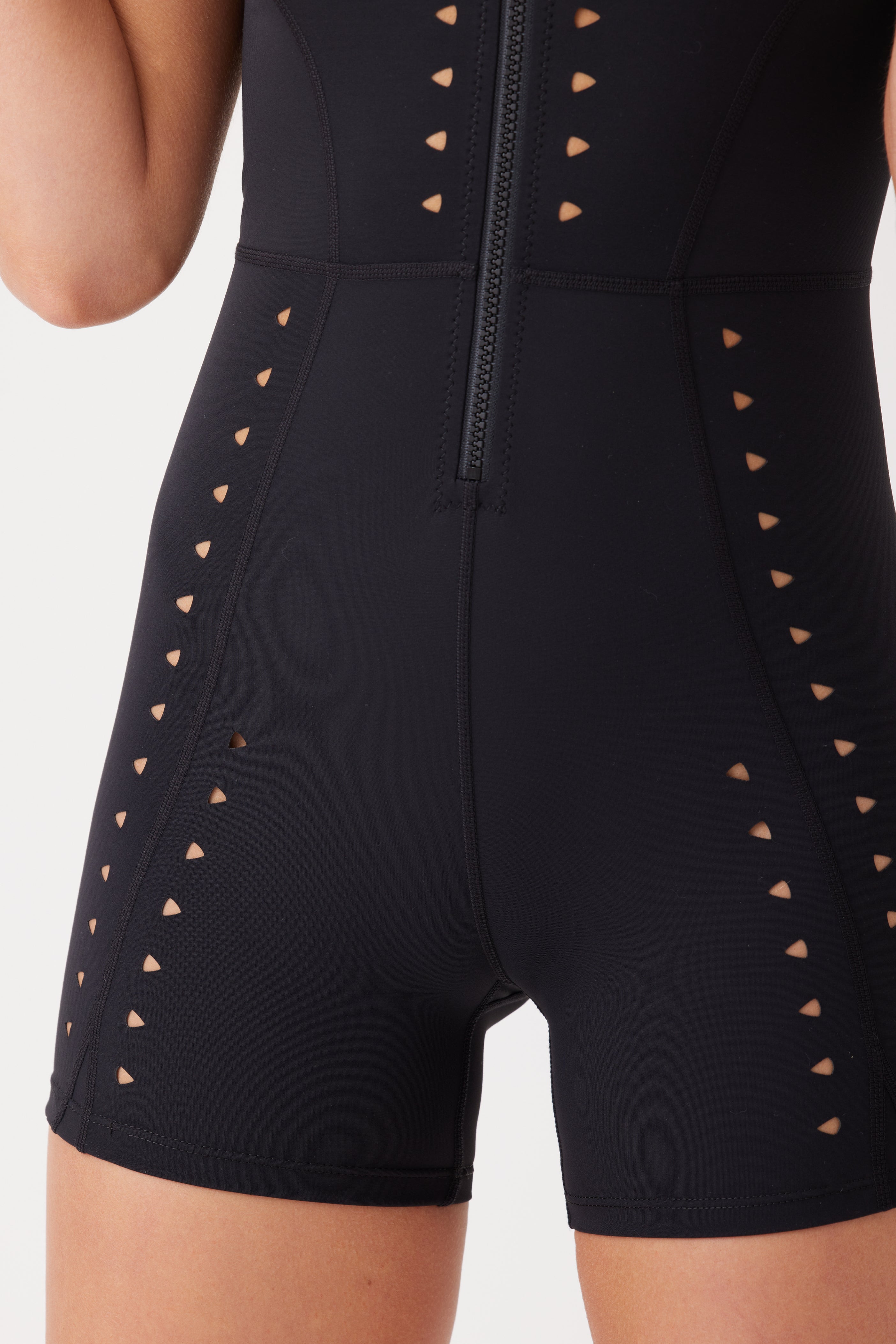 Close-up of a person in a black Kina Romper by Gigi C Sport, showcasing its laser cutout design with triangular patterns. This fitted garment features a front zipper and is crafted from breathable knit construction, set against a plain, light-colored background.