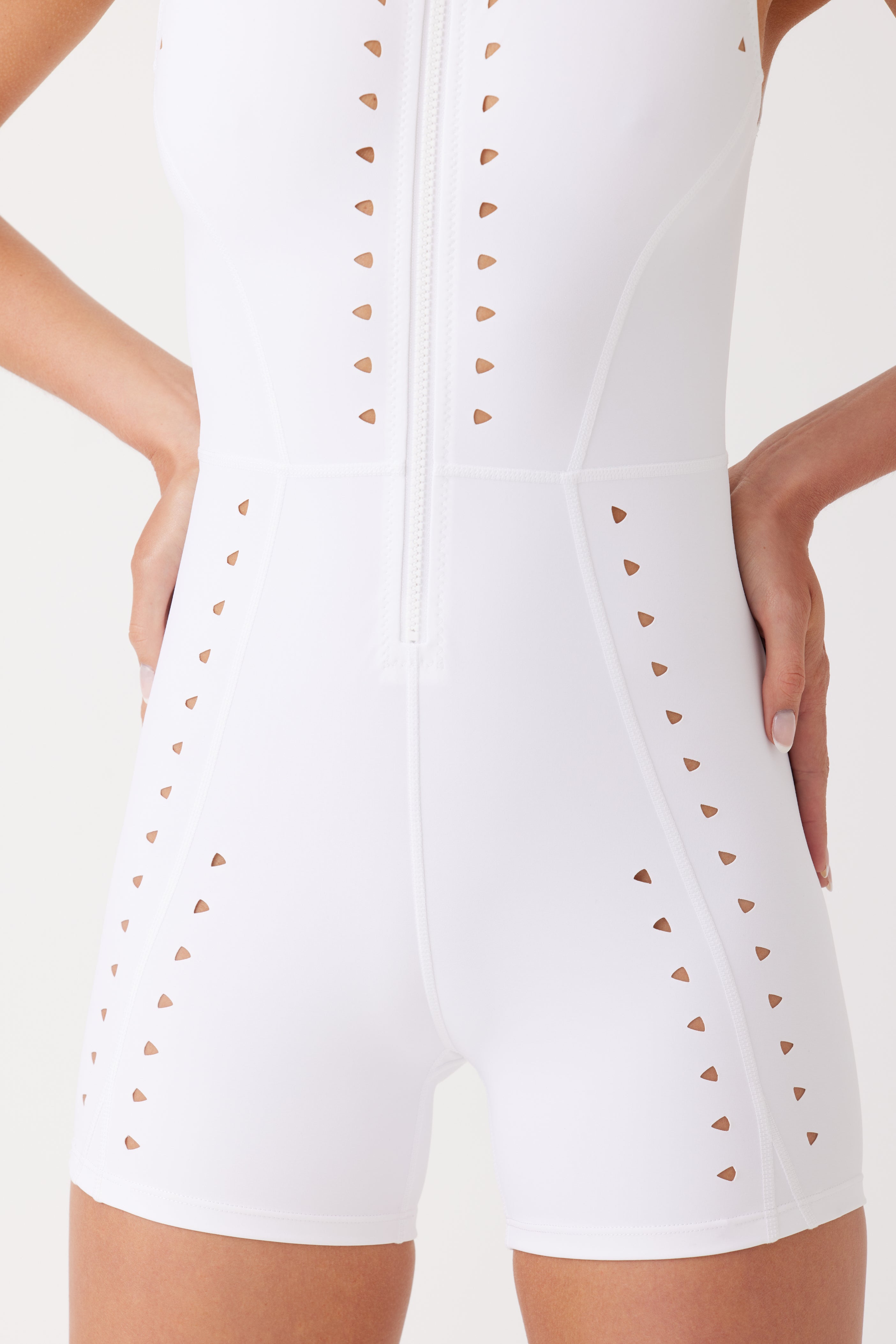 A person models the Gigi C Sports Kina Romper, a white, sleeveless outfit with triangle laser cutouts, a front zipper, and fitted design that highlights the torso and upper thighs, while their hands rest on their hips.