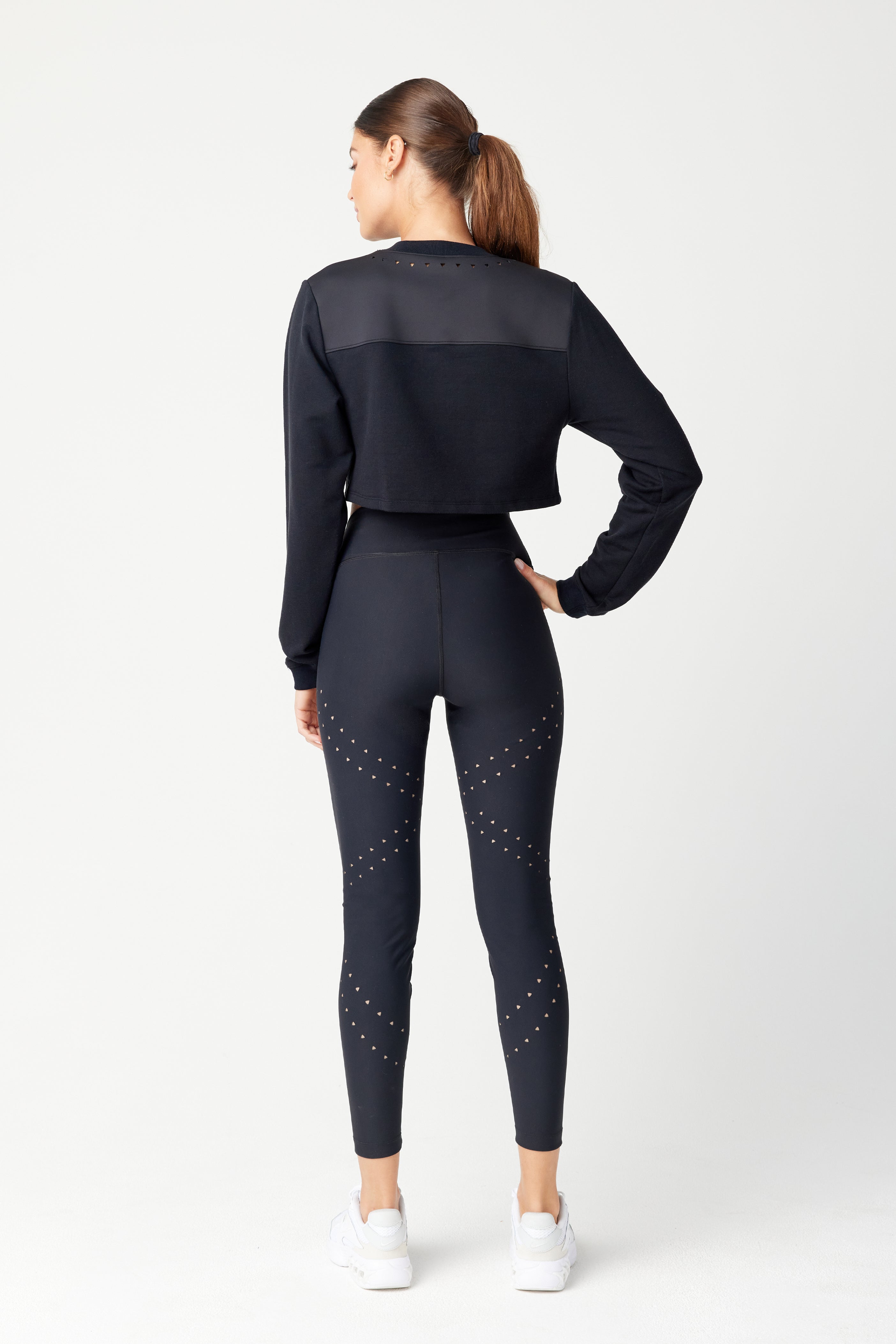 A woman with a ponytail stands with her back to the camera in Gigi C Sports chic Lina Pullover, made from recycled bonded Italian jersey. The tops triangle laser cutouts complement sleek leggings and white sneakers, all set against a light gray backdrop.