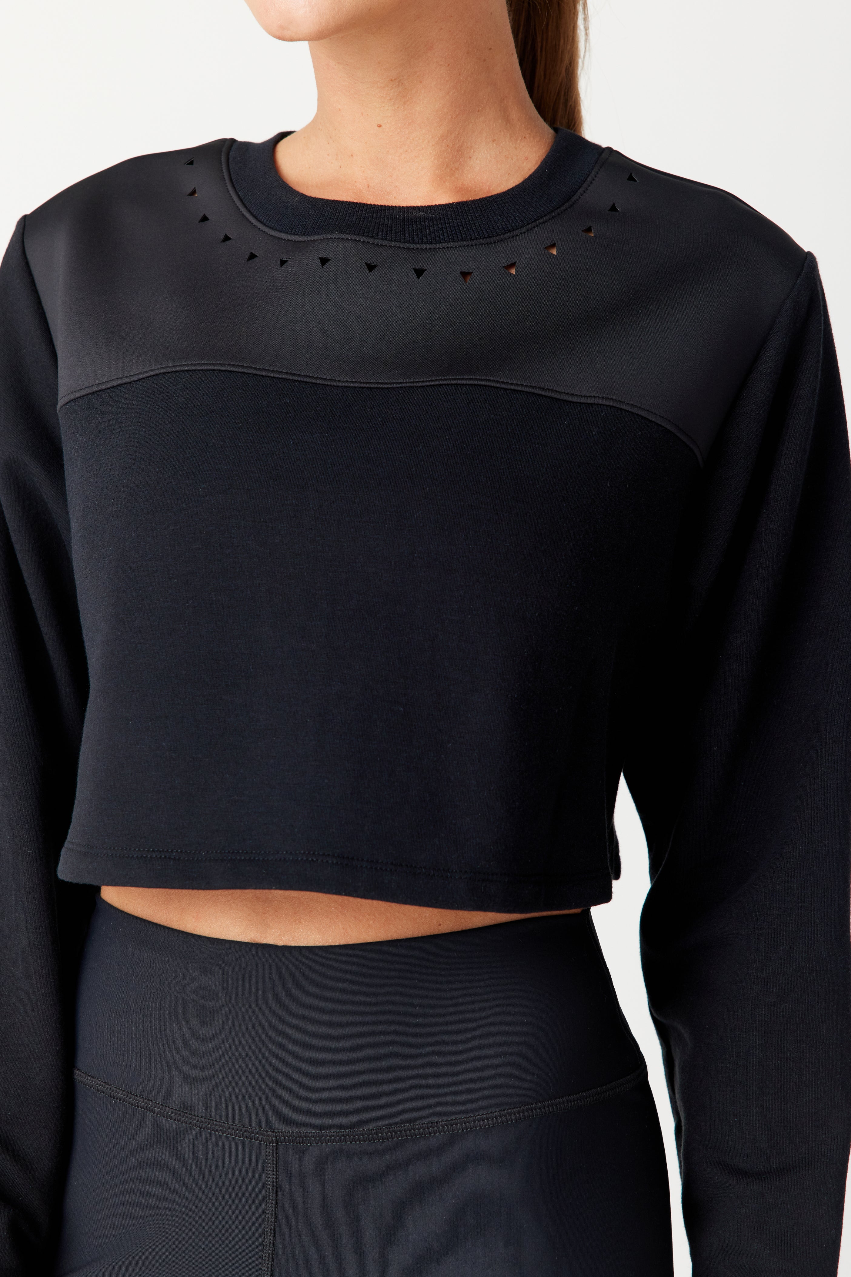 Someone is wearing a black Gigi C Sport Lina Pullover crafted from recycled bonded Italian jersey, with sophisticated triangle laser cutouts near the neckline. It’s paired with high-waisted black leggings against a plain off-white background.