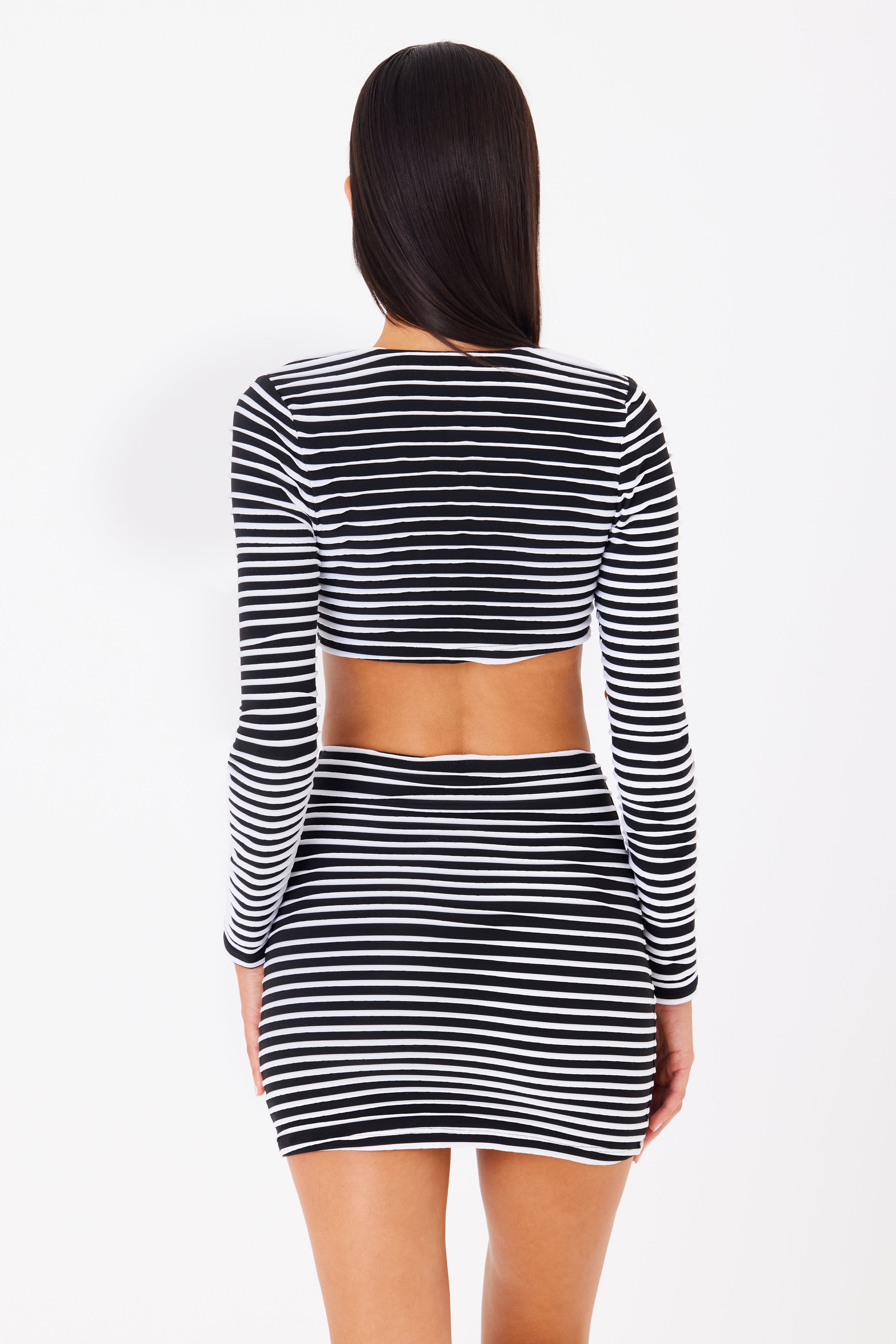 A person with long dark hair is stylishly dressed in the Lincoln Top by Gigi C Bikinis, featuring black and white stripes, long sleeves, a plunging neckline, and an open back. They pair it with a short skirt against a plain white backdrop.