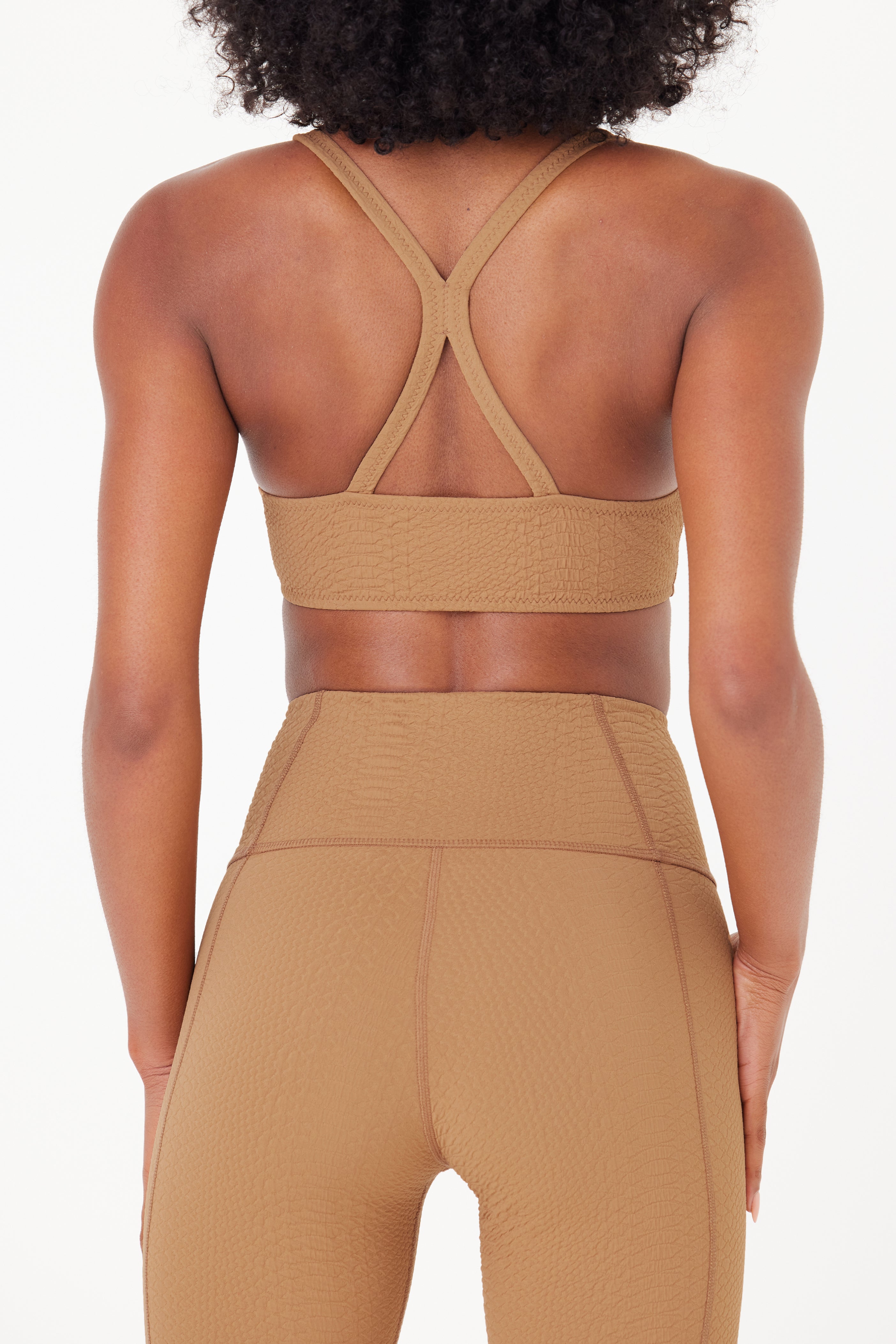 A person with curly hair is shown from the back, dressed in a tan bi-stretch compression athletic outfit. The look includes Gigi C Sports Marissa Bra, a textured medium-support sports bra with crossed straps, along with high-waisted leggings. The background is plain white.