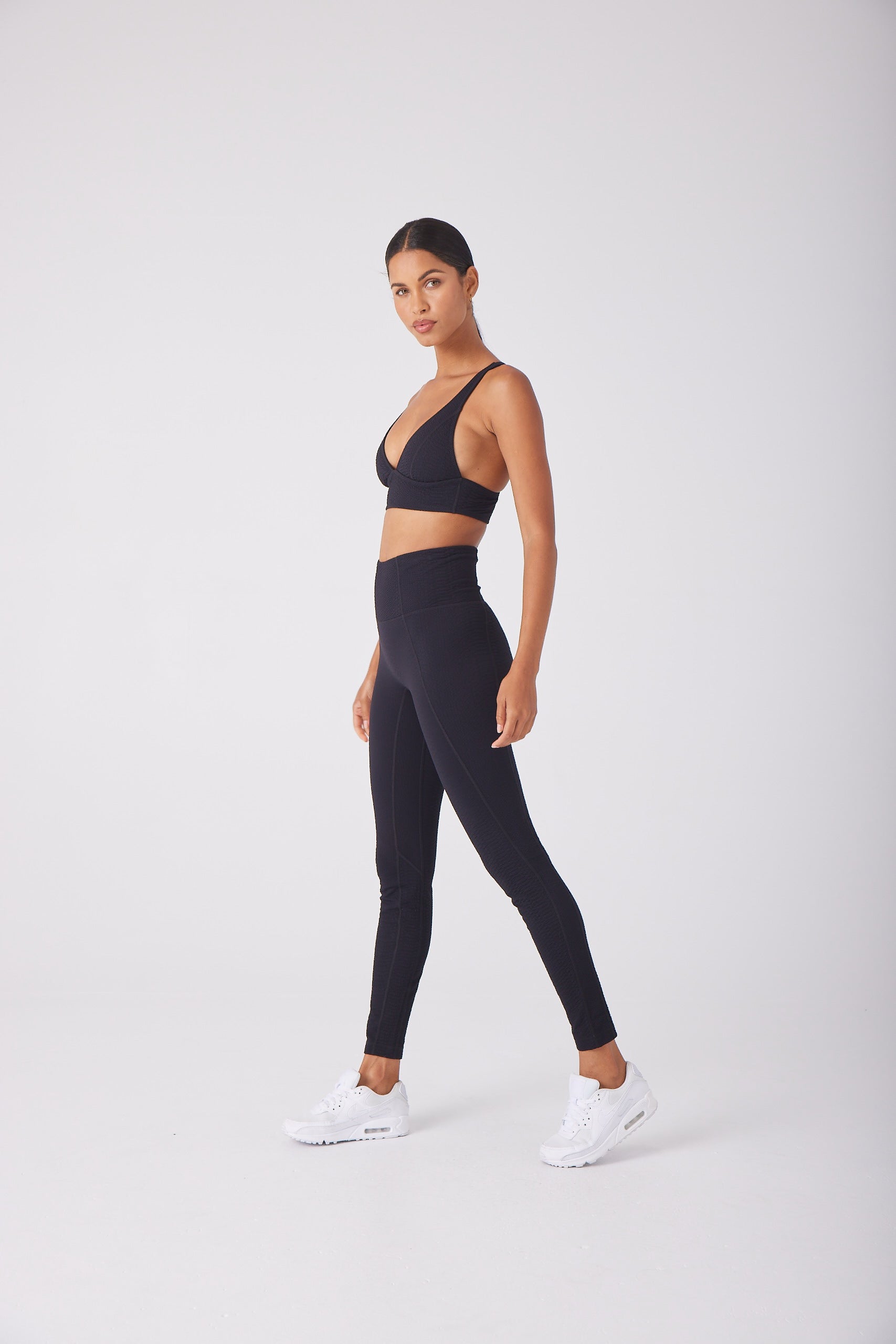 A person models the Marissa Bra from Gigi C Sport with leggings in black bi-stretch compression, standing neutrally against a white background while wearing white sneakers.