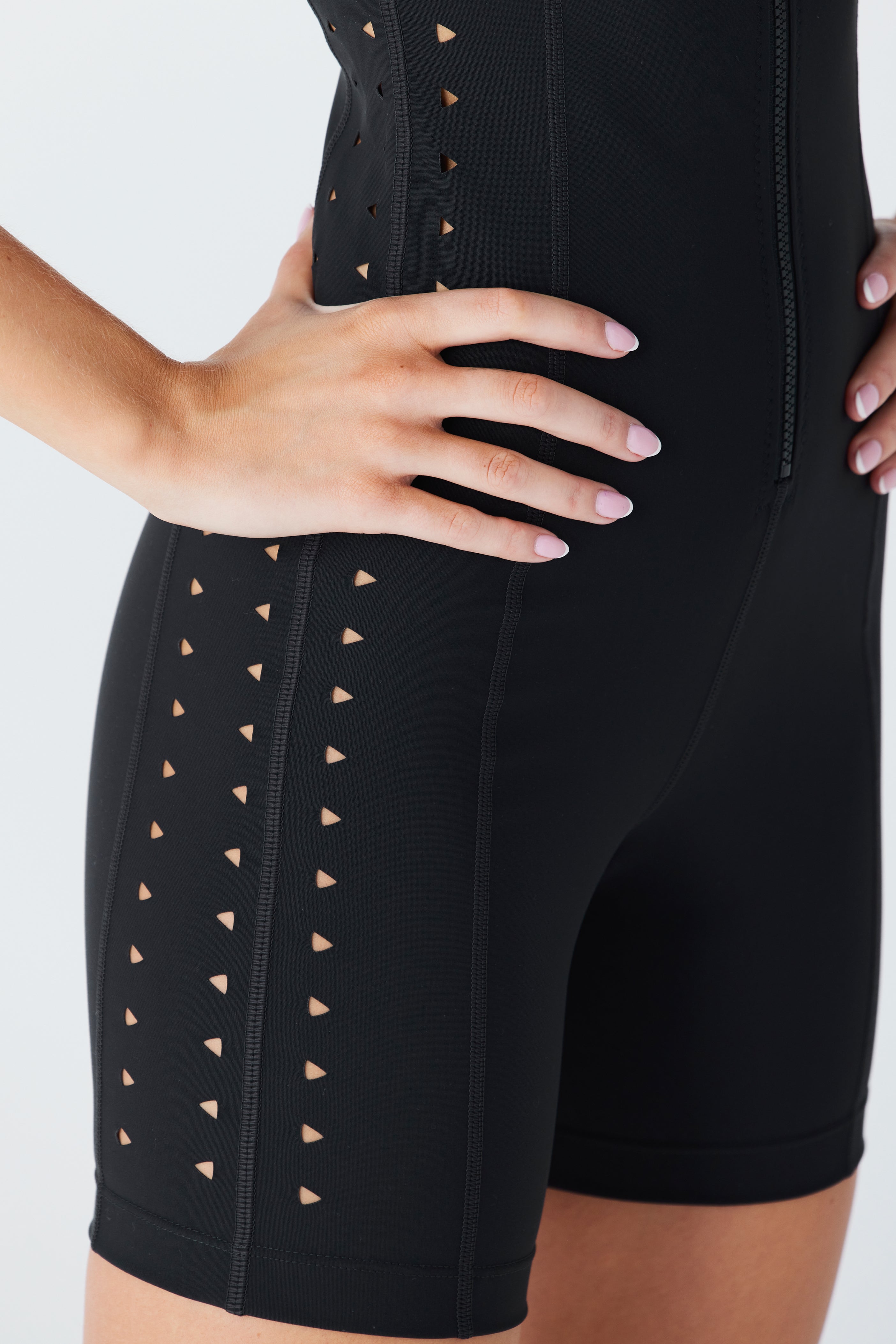 A person confidently poses with hands on hips in a stylish black Maxine Romper by Gigi C Sport, featuring advanced activewear fabric and small laser-cut triangular side cutouts, against a light-colored background.