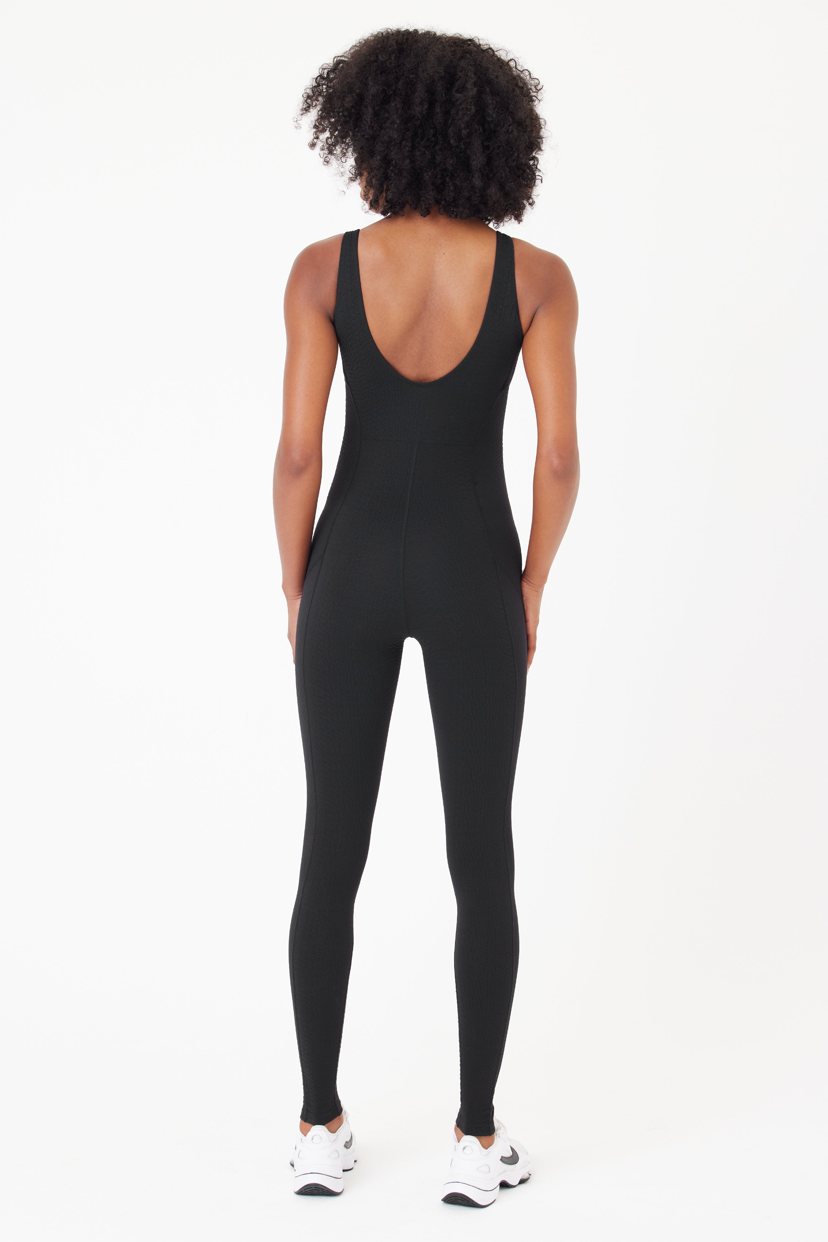 A person with curly hair, wearing the sleek black Merit Jumpsuit by Gigi C Sport featuring a bi-stretch compression fit and deep scoop back, along with white sneakers, is facing away.