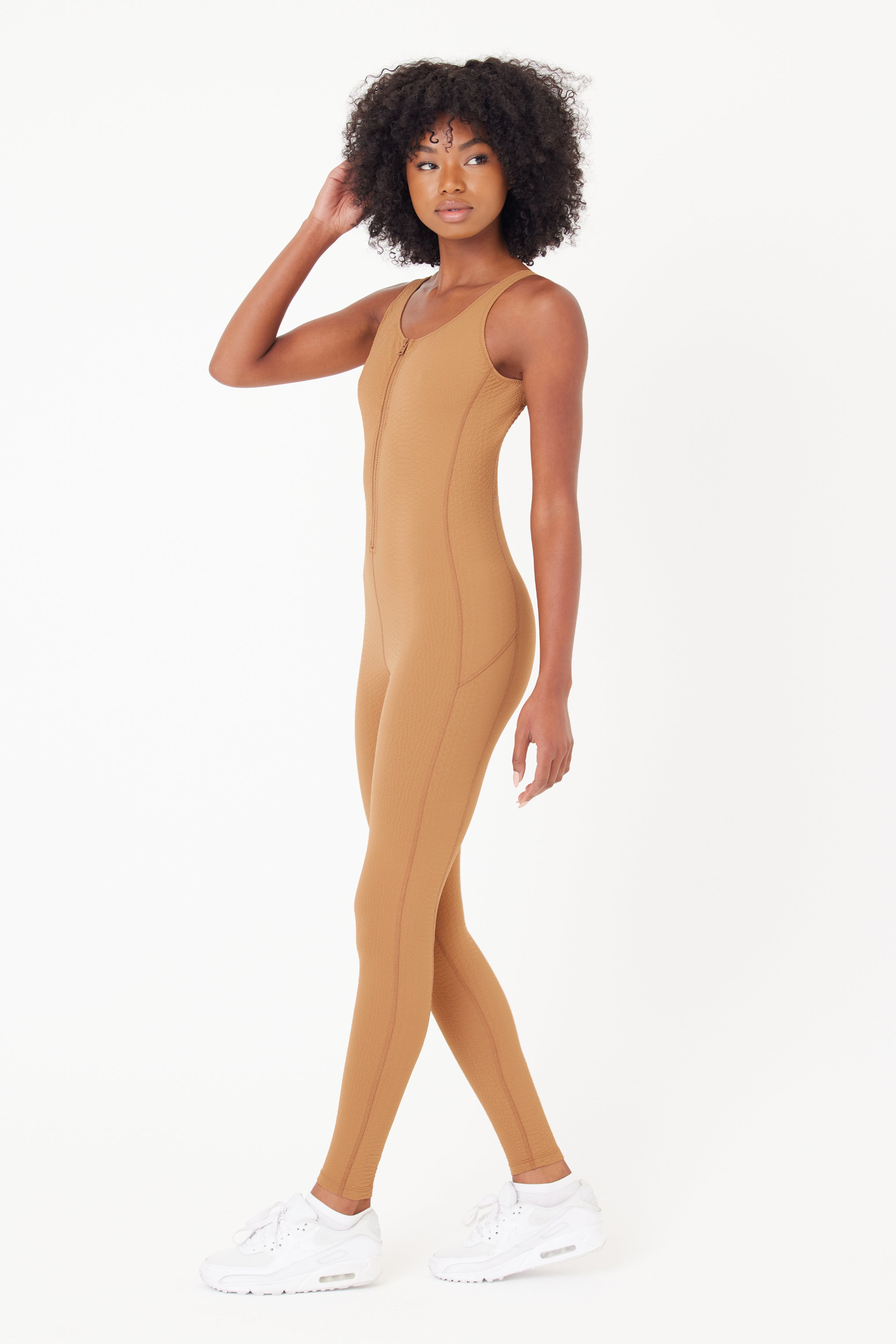 A person in the Gigi C Sport Merit Jumpsuit, a fitted tan outfit made from waffle jacquard fabric with thin shoulder straps, stands against a white background. They touch their curly hair while glancing to the side, wearing sleek white sneakers.