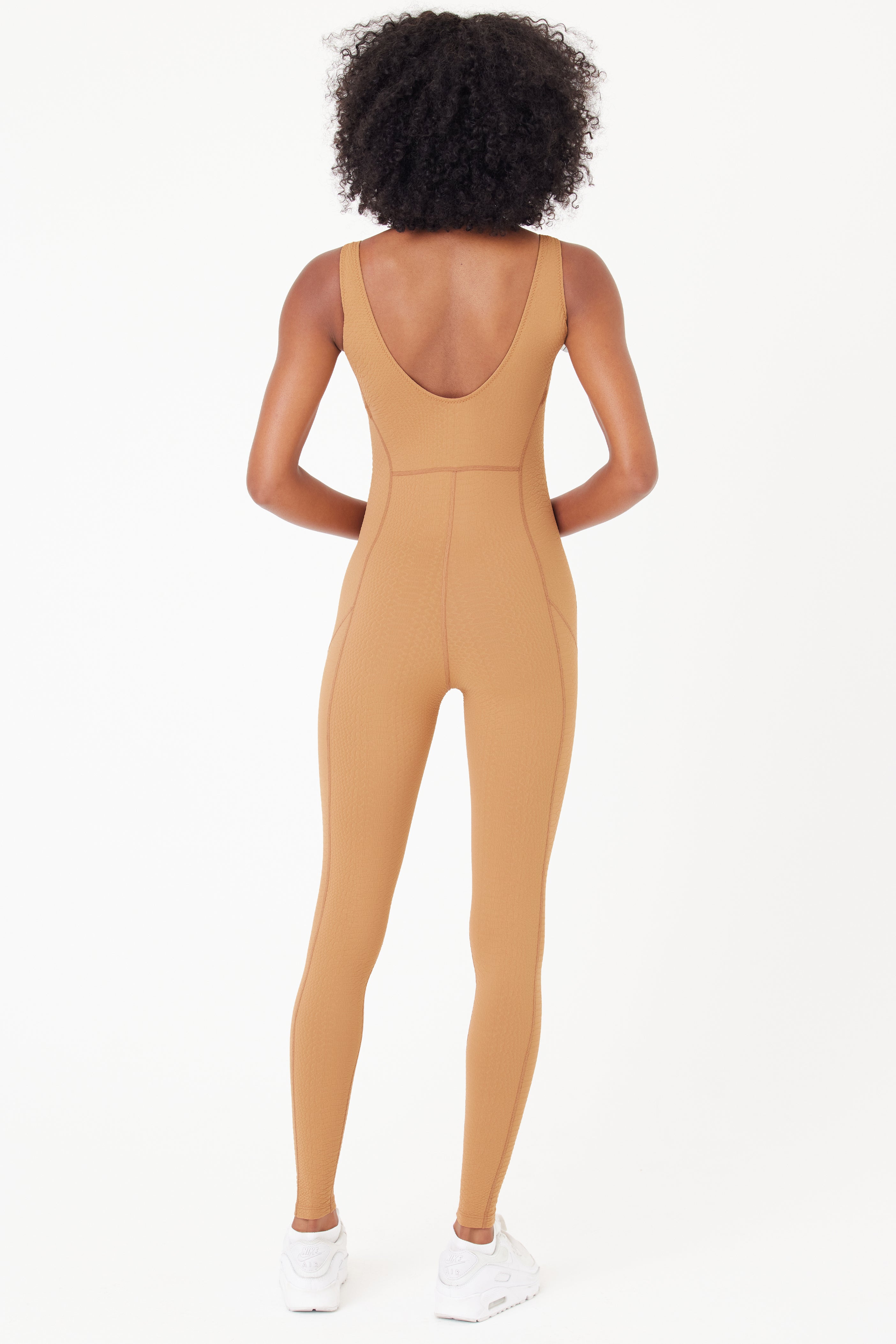 A person with curly hair, barefoot and facing away against a white background, is wearing the Gigi C Sport Merit Jumpsuit—a tan, sleeveless, full-length fitted outfit made from waffle jacquard fabric.