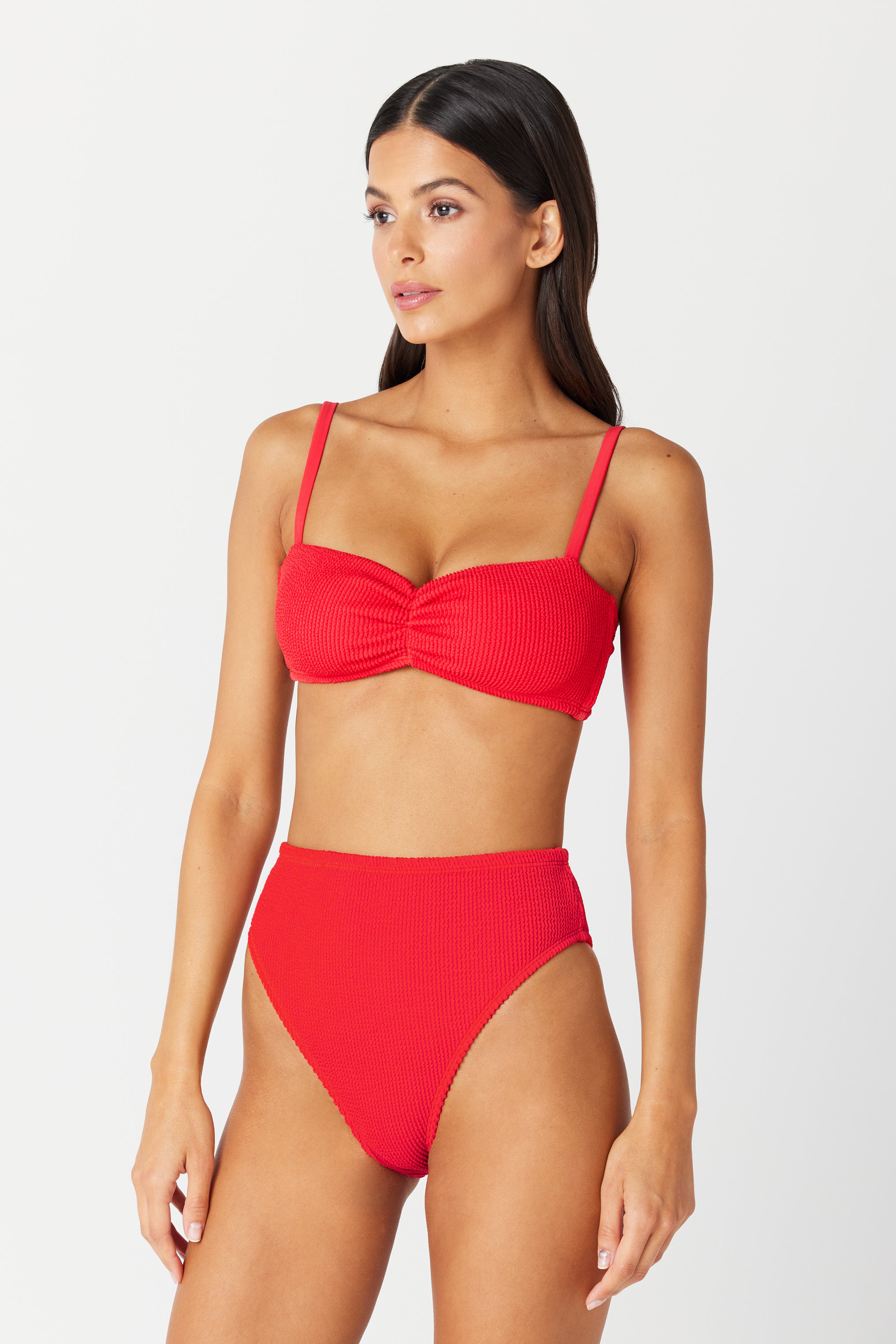 A person with long dark hair is wearing a red Malia Bottom from Gigi C Bikinis, featuring cheeky coverage and an ultra-high cut leg. The top has thin shoulder straps and a ruched front. The background is plain and light-colored.