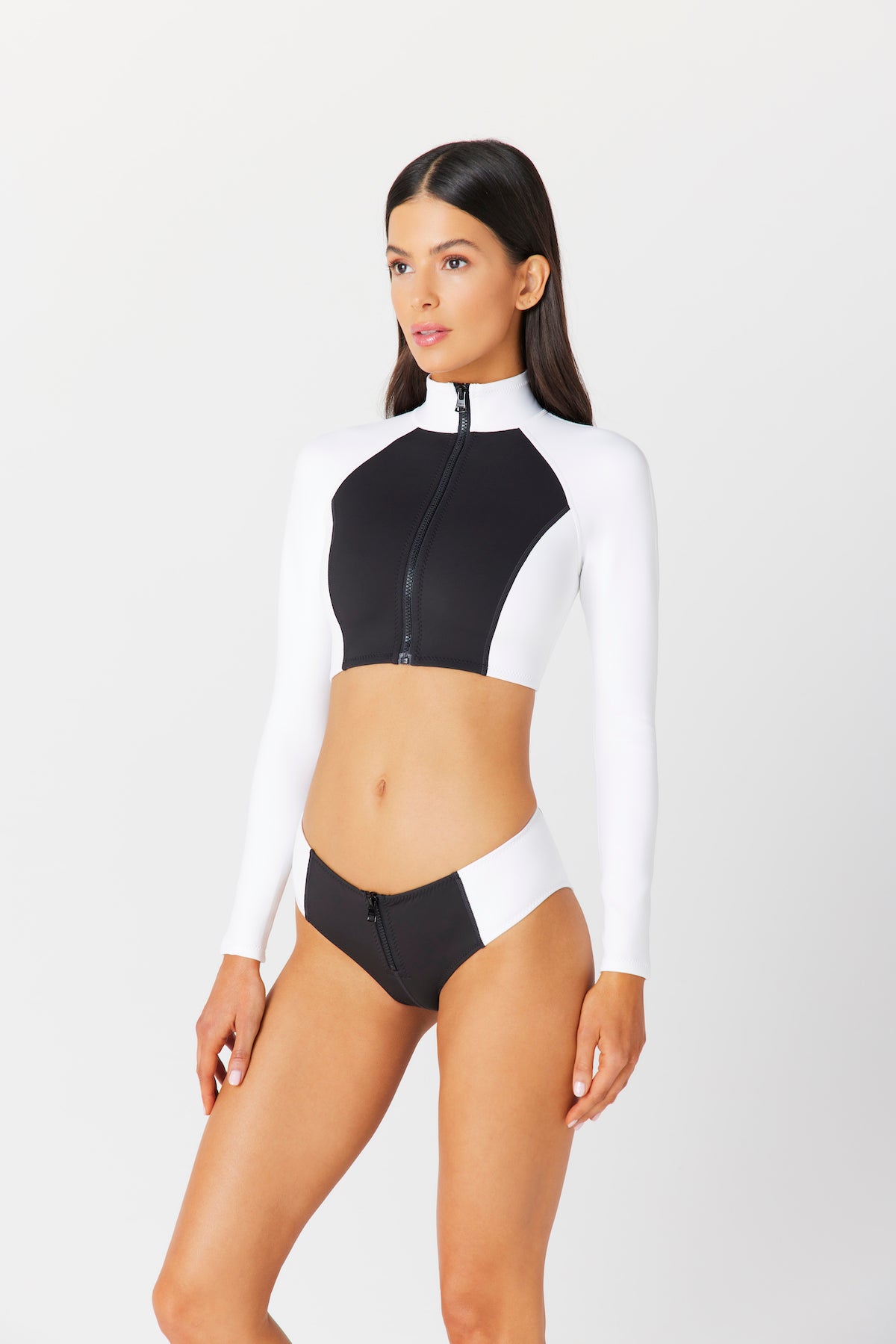 A woman models a black and white long-sleeve crop top with a zipper and Bianca Short by Gigi C Bikinis, standing against a plain white background and gazing slightly to the side.