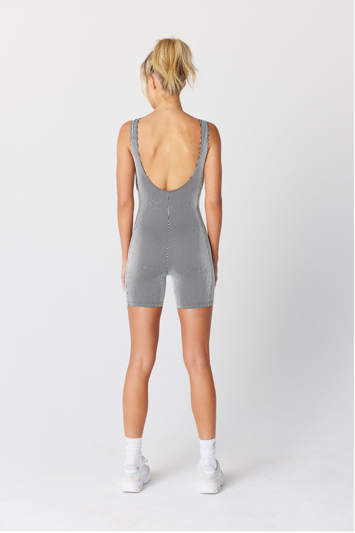 A person, with blonde hair in a ponytail, is seen from behind wearing the gray Monti Romper by Gigi C Sport. The sleeveless textured romper is paired with white sneakers on a plain white background.