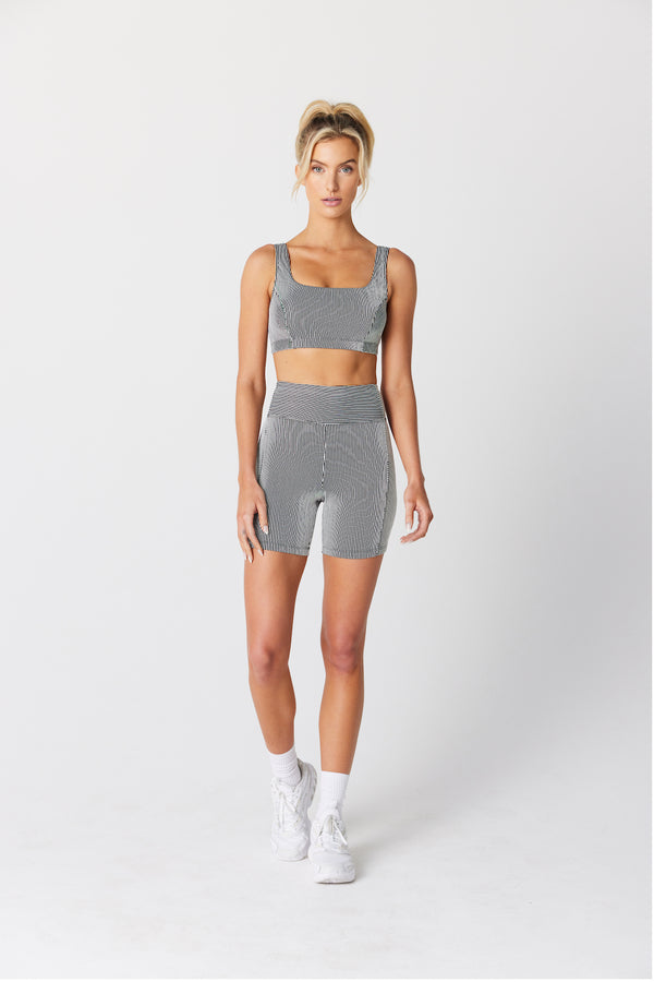 Jess Short models a gray sports bra and shorts with mini stripes from Gigi C Sport, paired with white socks and shoes. Her hair is tied up as she confidently faces the camera against a plain background.