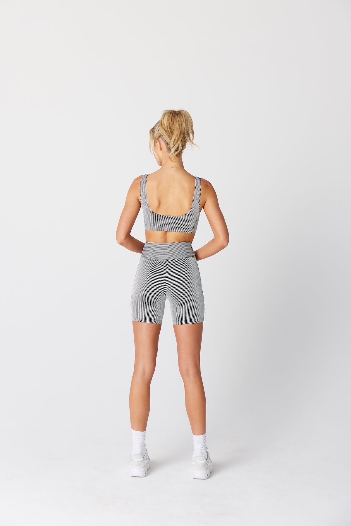 A blonde-haired person in a ponytail wears a grey sports top, Gigi C Sports Jess Short, white socks, and sneakers. They pose with their hands behind their back against a plain white background.