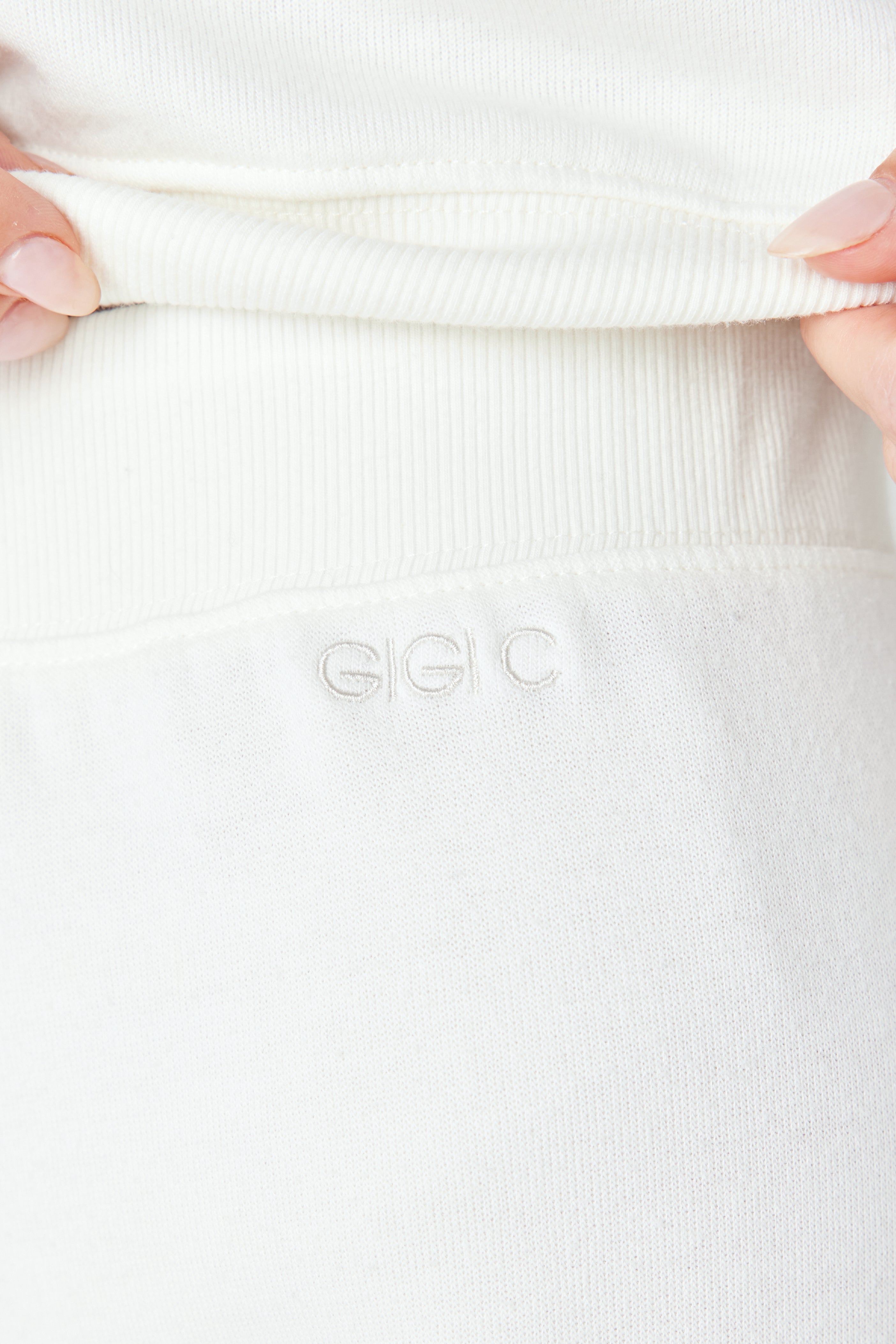 A close-up shows neatly manicured hands gently pulling the waistband of a white garment, echoing the sleek design of the Gigi C Lounges Harlow Pant. The fabric is adorned with GIGI C stitching.