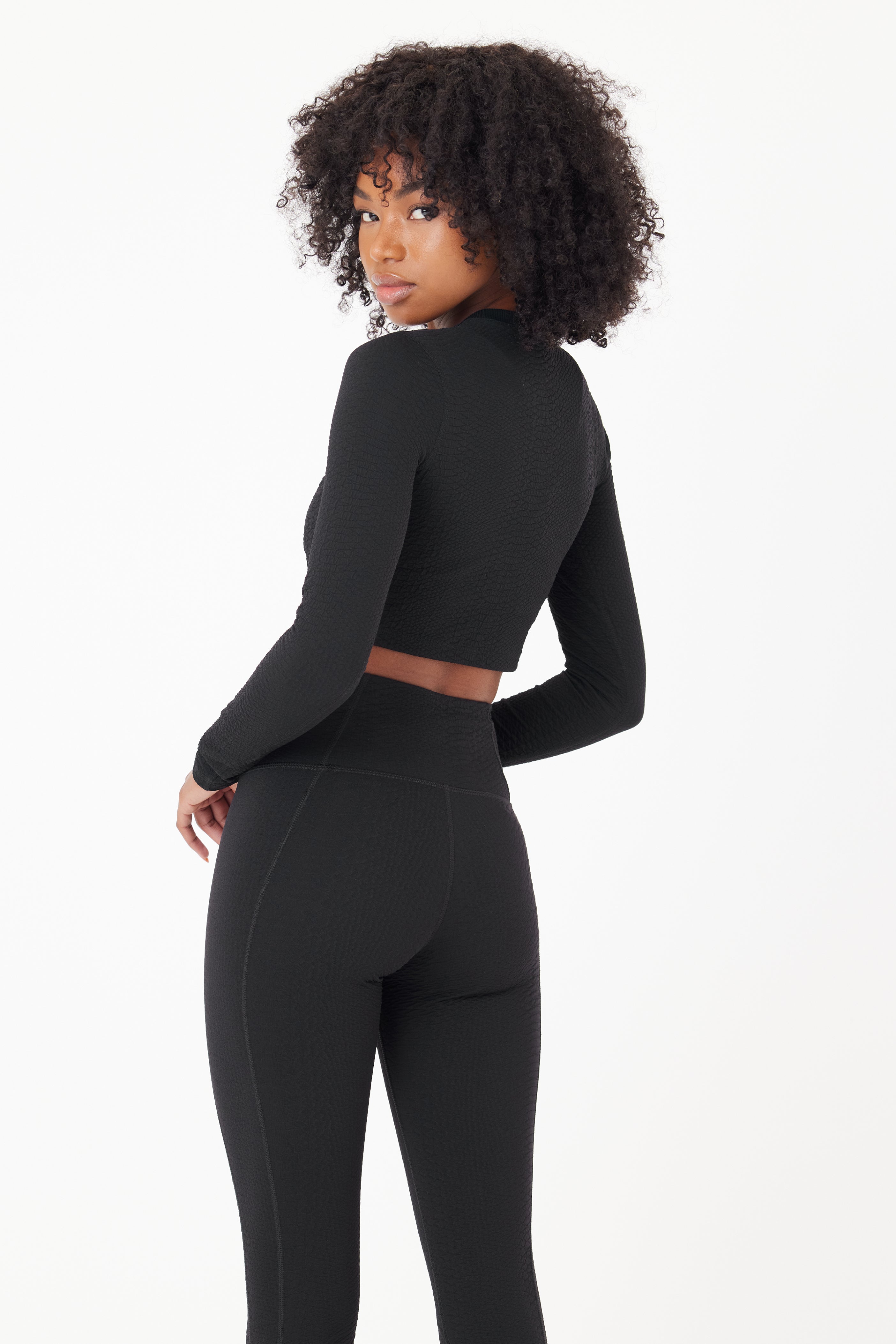 A person with curly hair wearing a Gigi C Sport Quinn Top and matching Ava Legging, both textured to resemble faux snake skin, looks over their shoulder against a plain light background.
