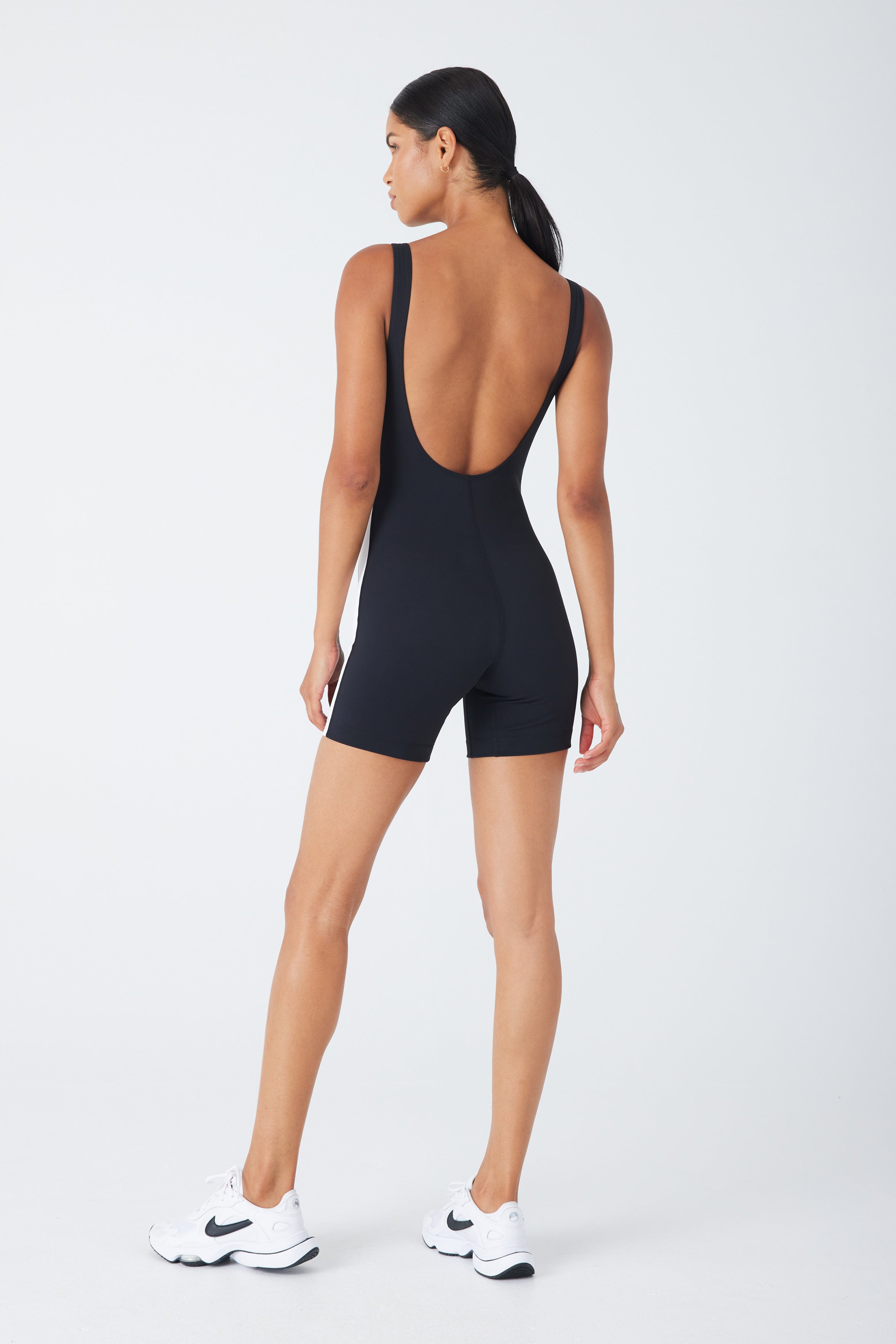 A person faces away from the camera wearing the Theo Romper by Gigi C Sport. The chic black, sleeveless design with a low back pairs seamlessly with white athletic shoes, set against a plain white background.